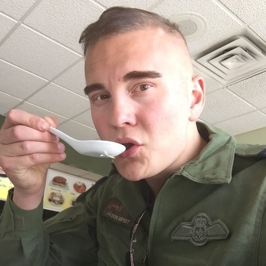Young man with short hair and light skin holding a spoon near his mouth, wearing a green jacket with patches.