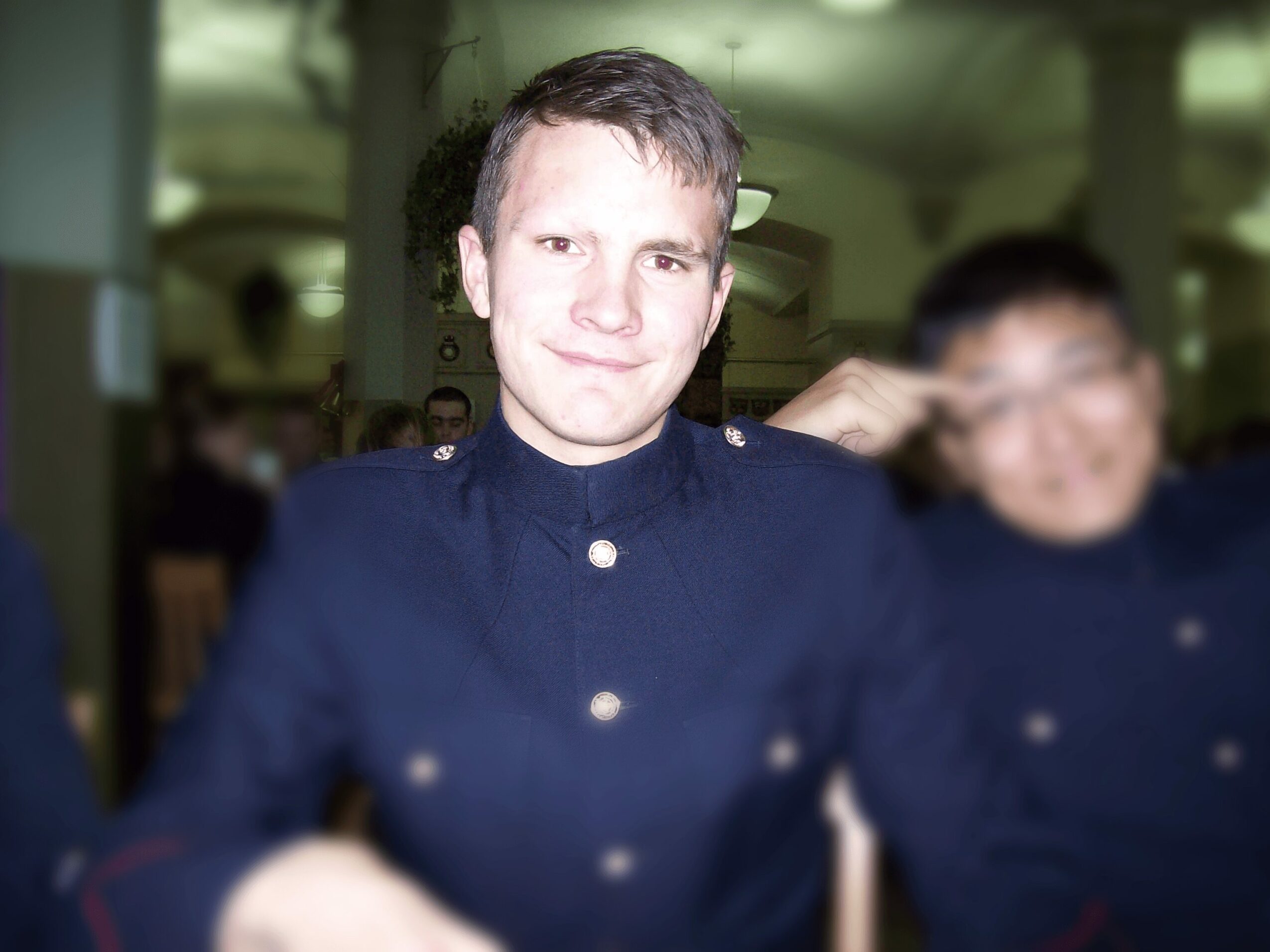 Young man in a dark uniform smiling, with blurred person beside him, indoors with columns in background.