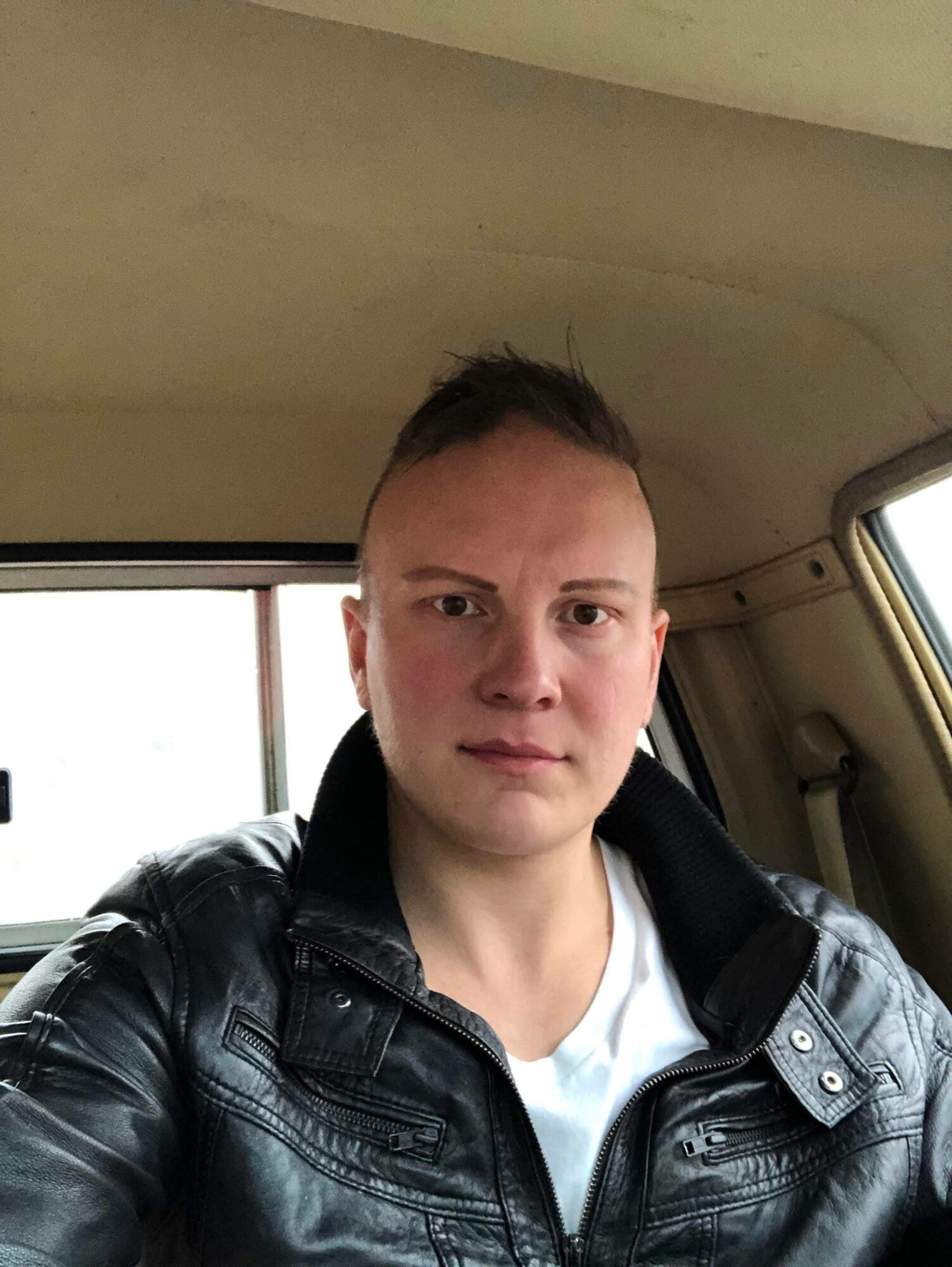 Person with short hair wearing a black leather jacket inside a vehicle, looking at the camera.