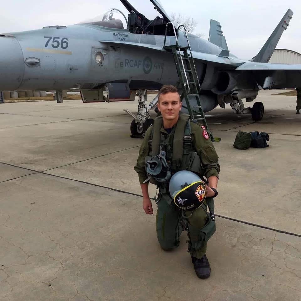 Kneeling pilot in uniform holding helmet with aircraft and jet in background.