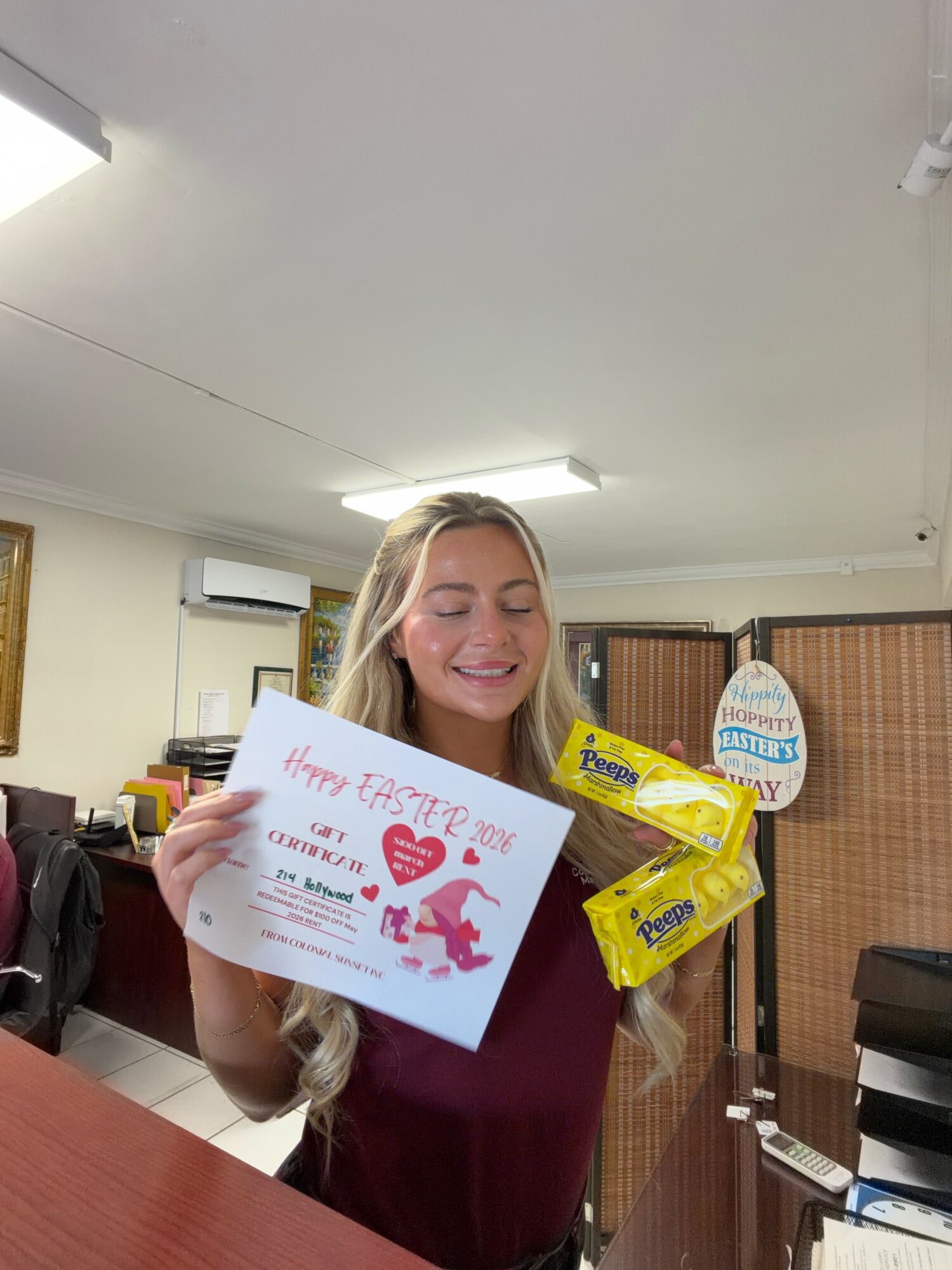 Young woman with long blonde hair smiling, holding a colorful Easter card and a yellow snack package indoors.