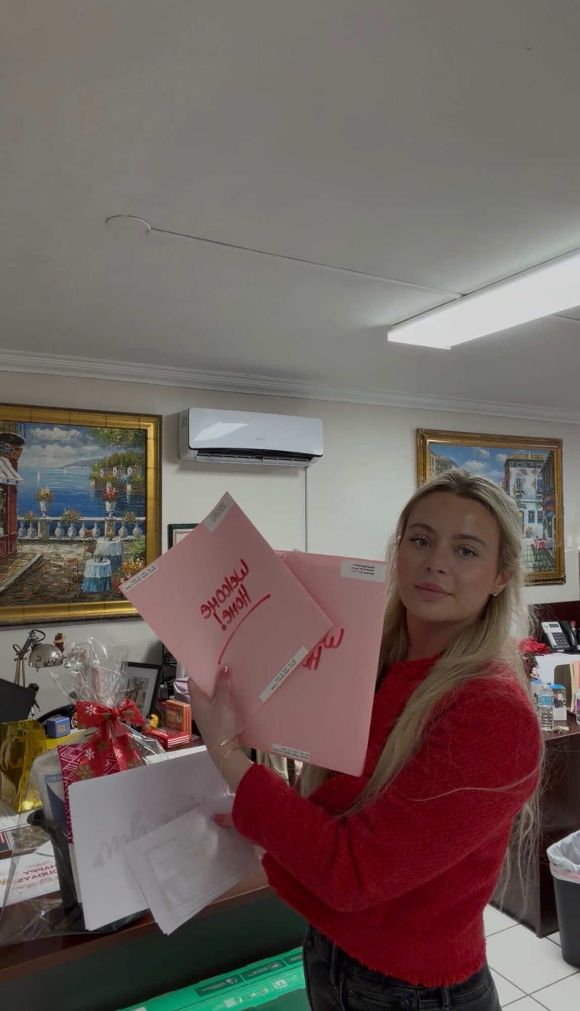 Young woman with long blonde hair holding pink folders in a room with paintings and office supplies.