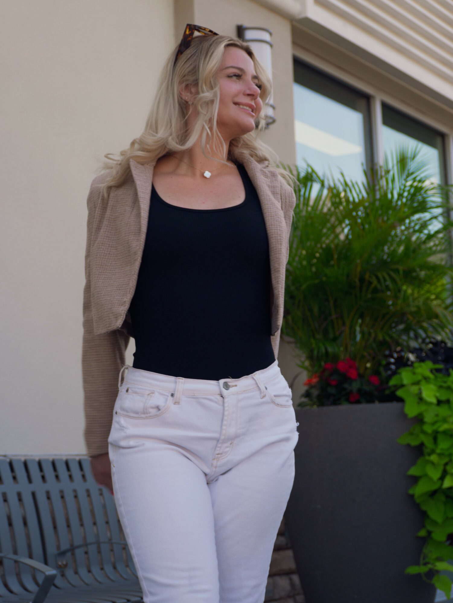 Woman with blonde hair smiling outdoors, wearing a black top, beige cardigan, and white pants, near plants and a building.