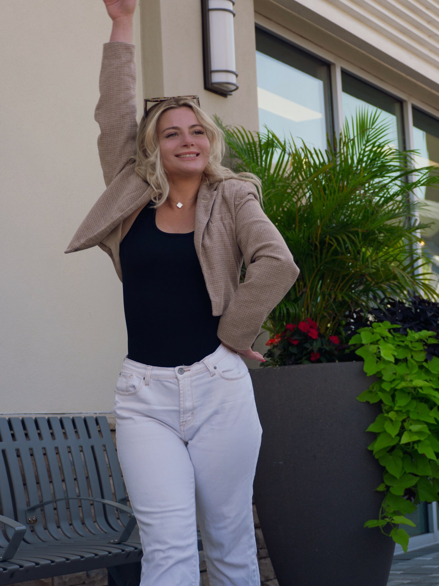 Woman smiling outdoors, raising her arm, standing next to a large plant and a building window.
