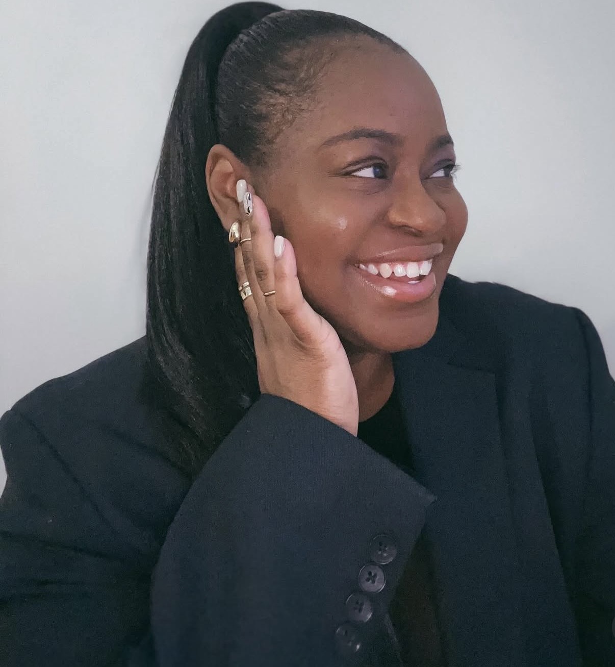 Smiling woman with long hair in a ponytail, wearing a dark blazer, touching her face with her hand.
