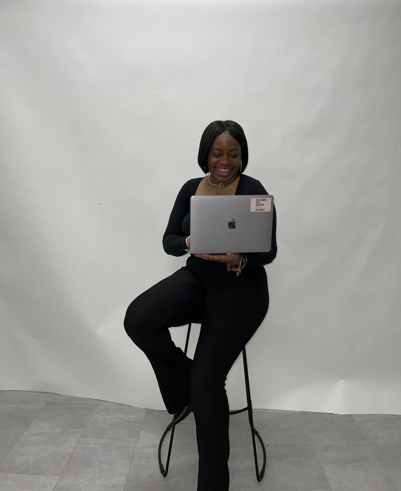 Woman sitting on a stool, using a laptop, smiling, in front of a plain white background.