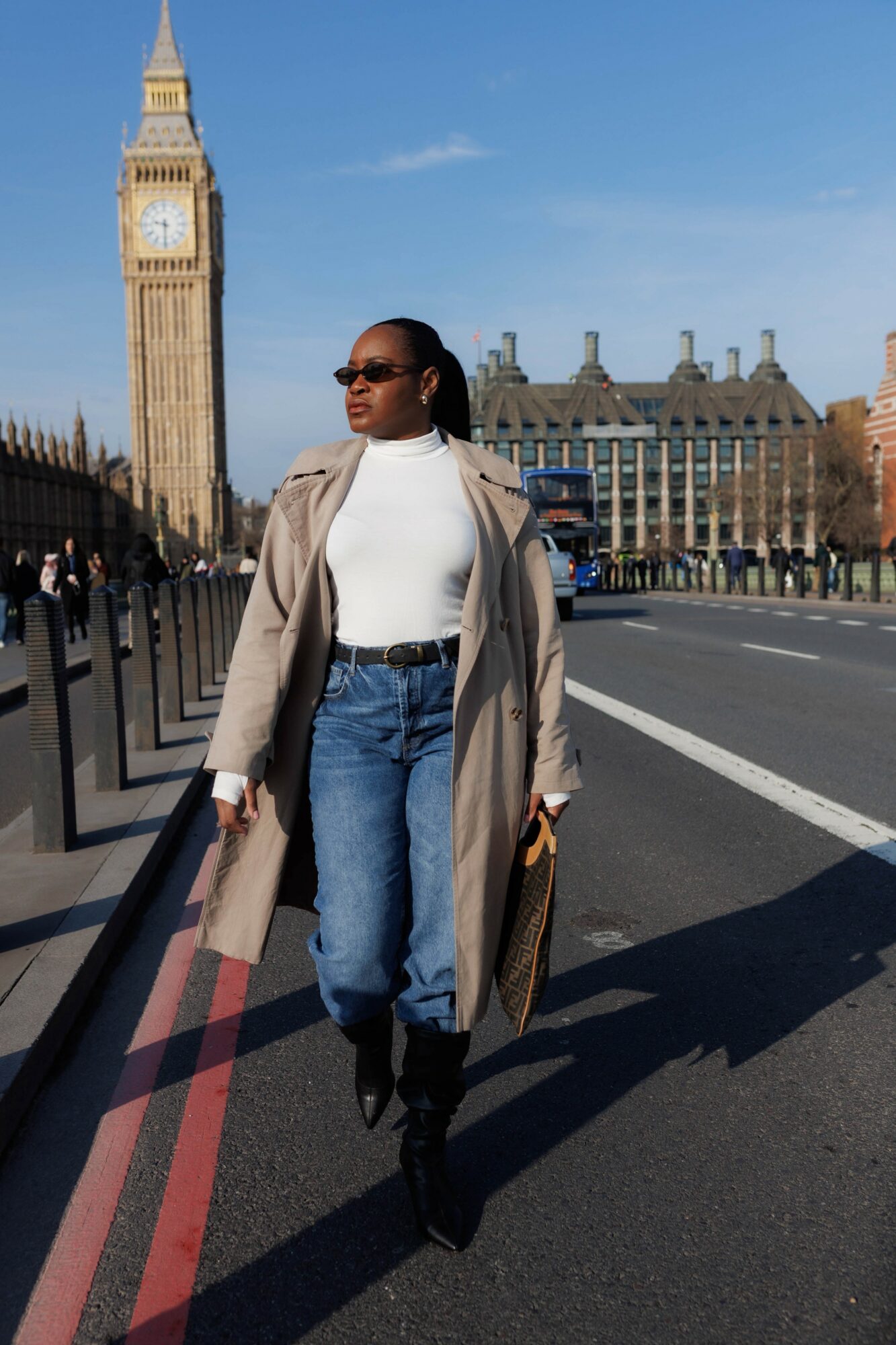 Woman walking on street with Big Ben in background, wearing sunglasses, coat, white top, and jeans.