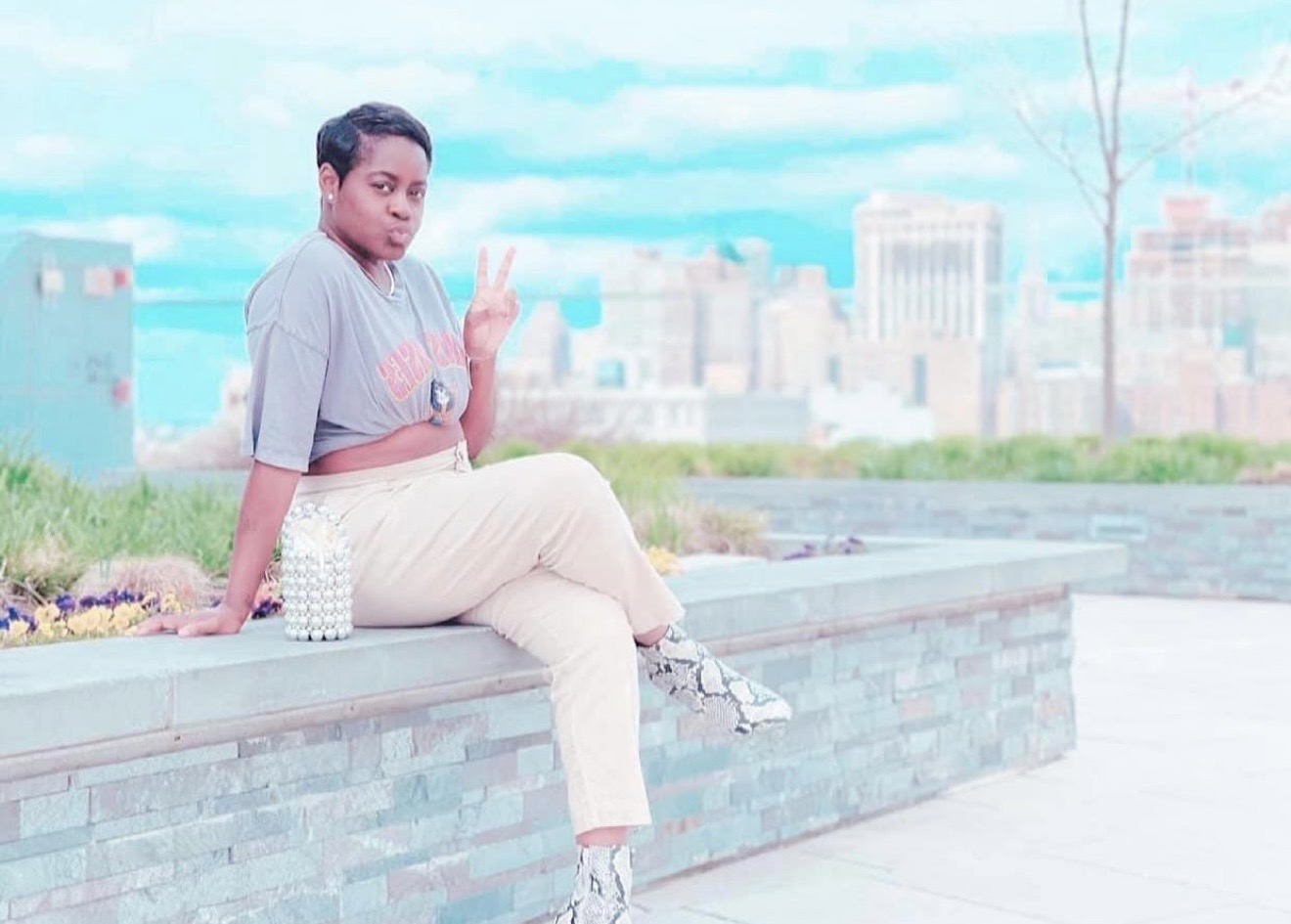 Person sitting on a brick ledge outdoors, cityscape in background, making a peace sign with hand.