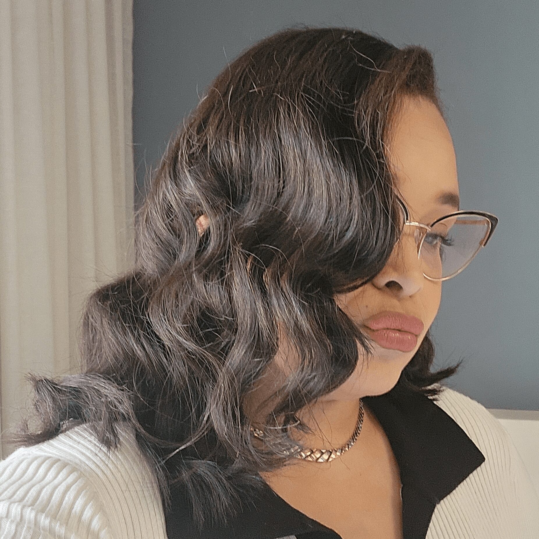 Woman with wavy dark hair, glasses, and a necklace, looking to the side indoors.