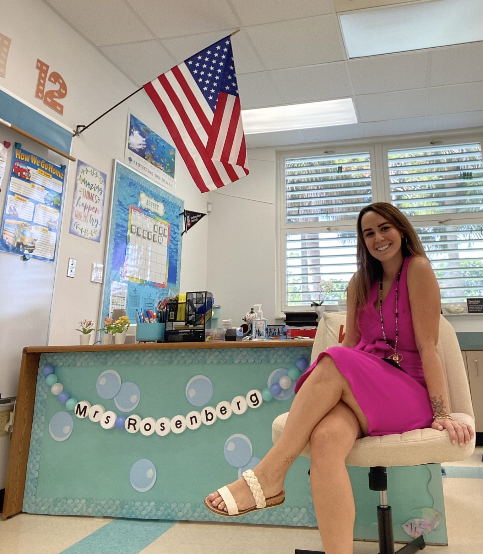 Woman in pink dress sitting on chair in classroom with American flag and decorated desk.