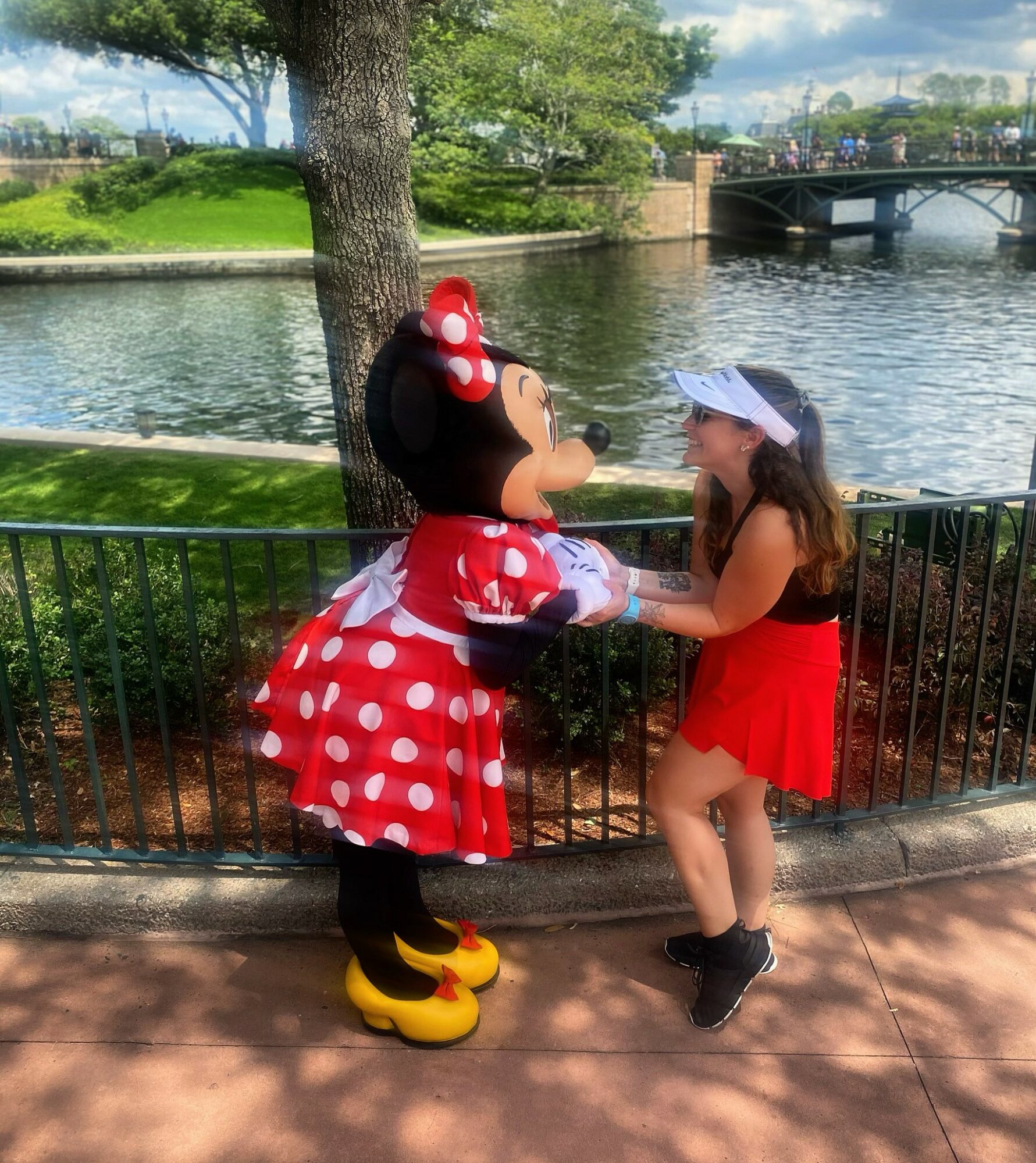 Girl in red skirt and white cap holding hands with Minnie Mouse mascot near river, trees, and bridge in background.