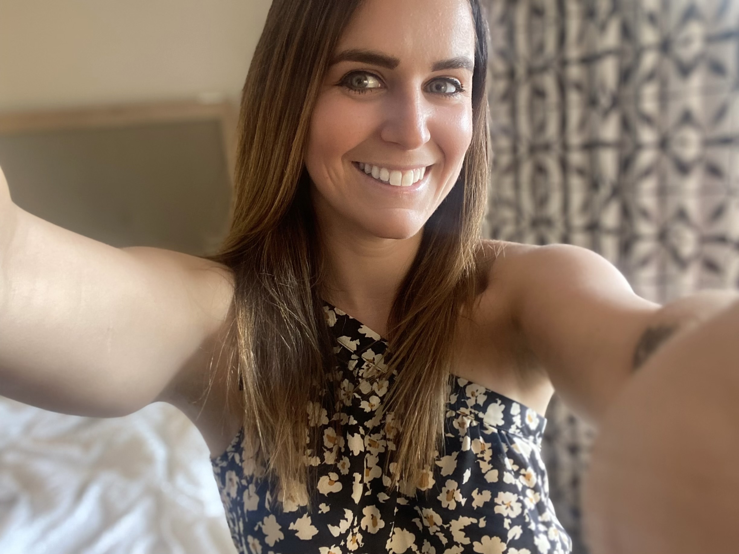 Woman smiling, taking a selfie indoors, wearing a floral top, with curtains in the background.