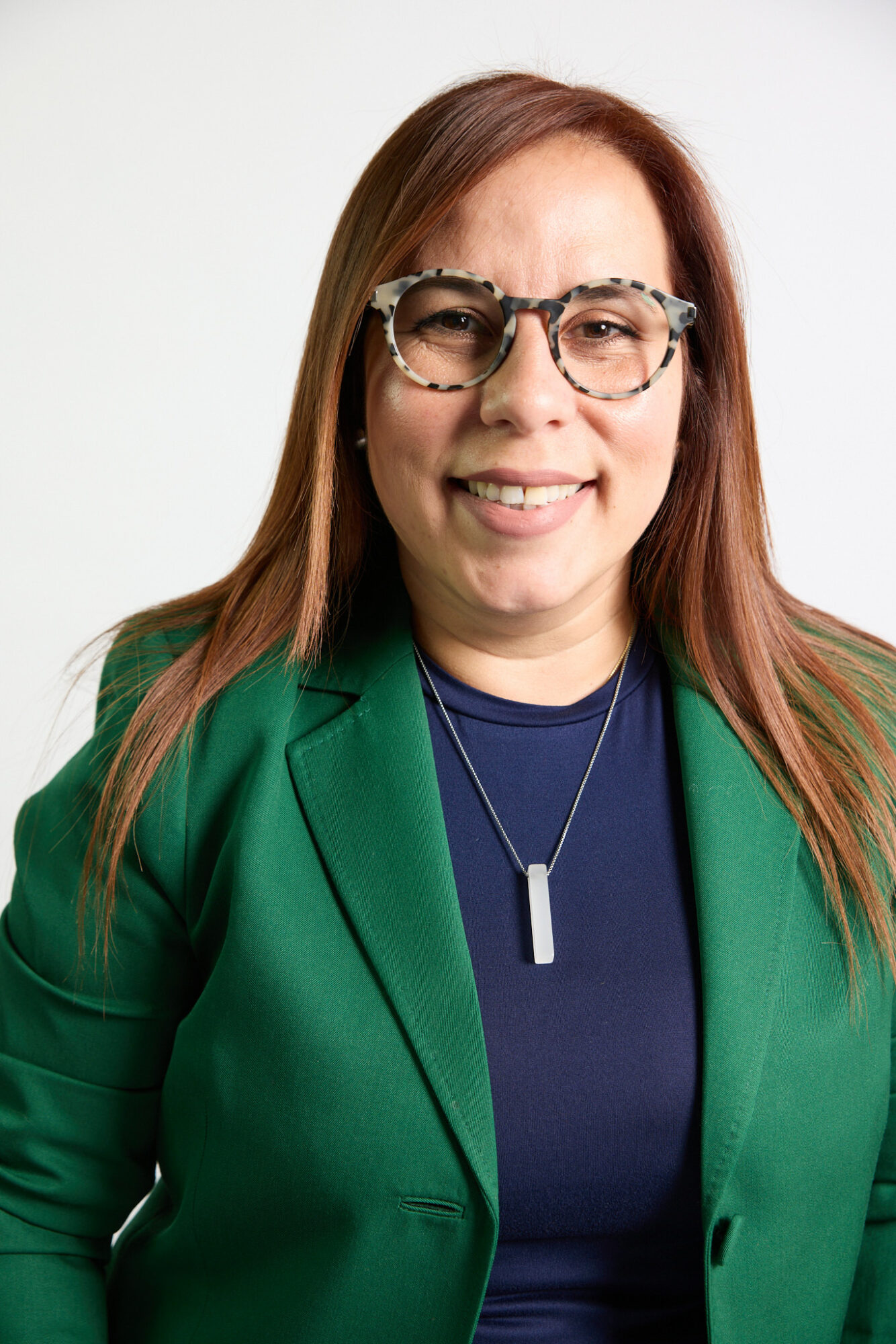 Woman with long brown hair wearing glasses, green blazer, and navy top, smiling at camera.