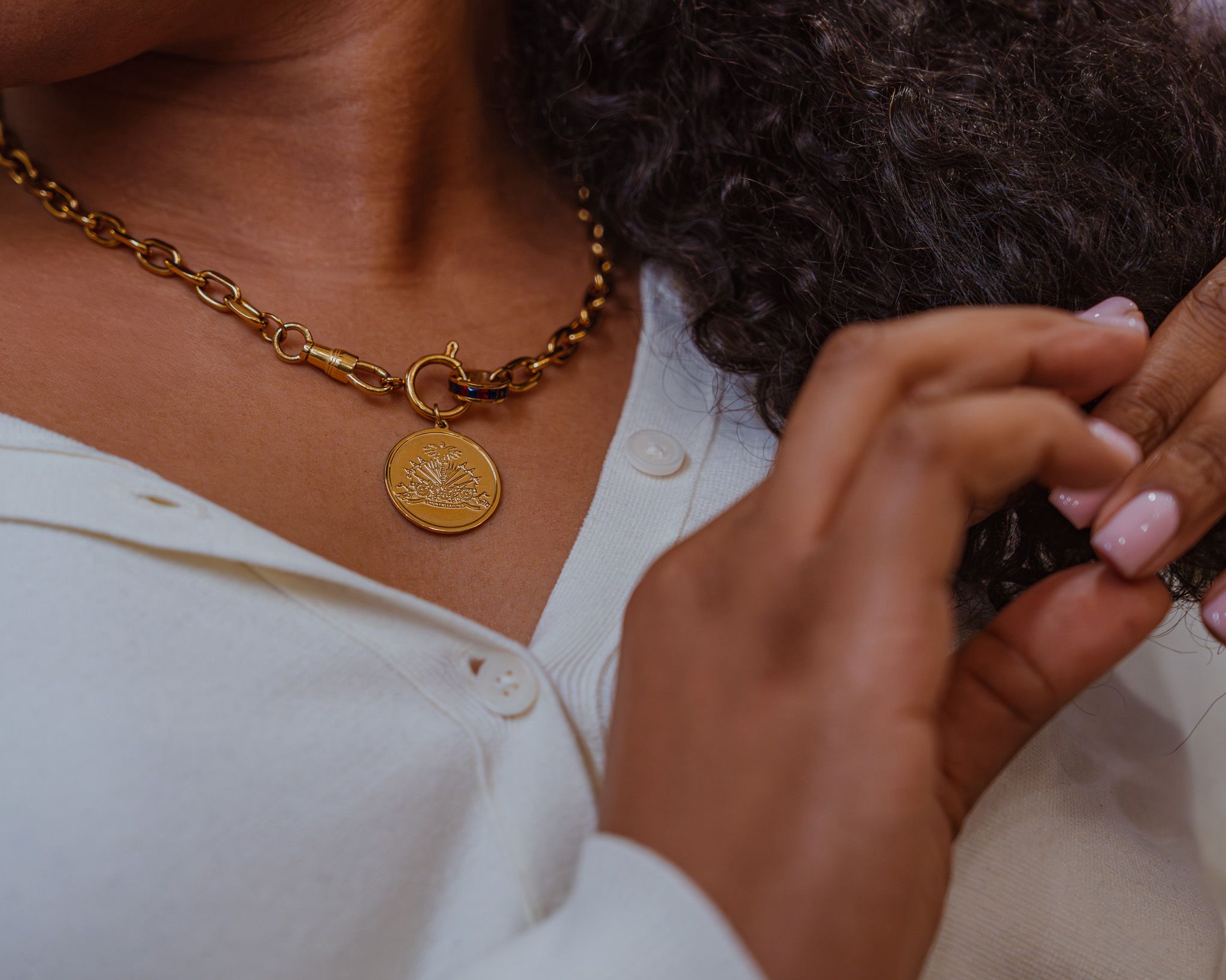 Close-up of a person wearing a gold chain necklace with a round pendant, white shirt, and hand near hair.