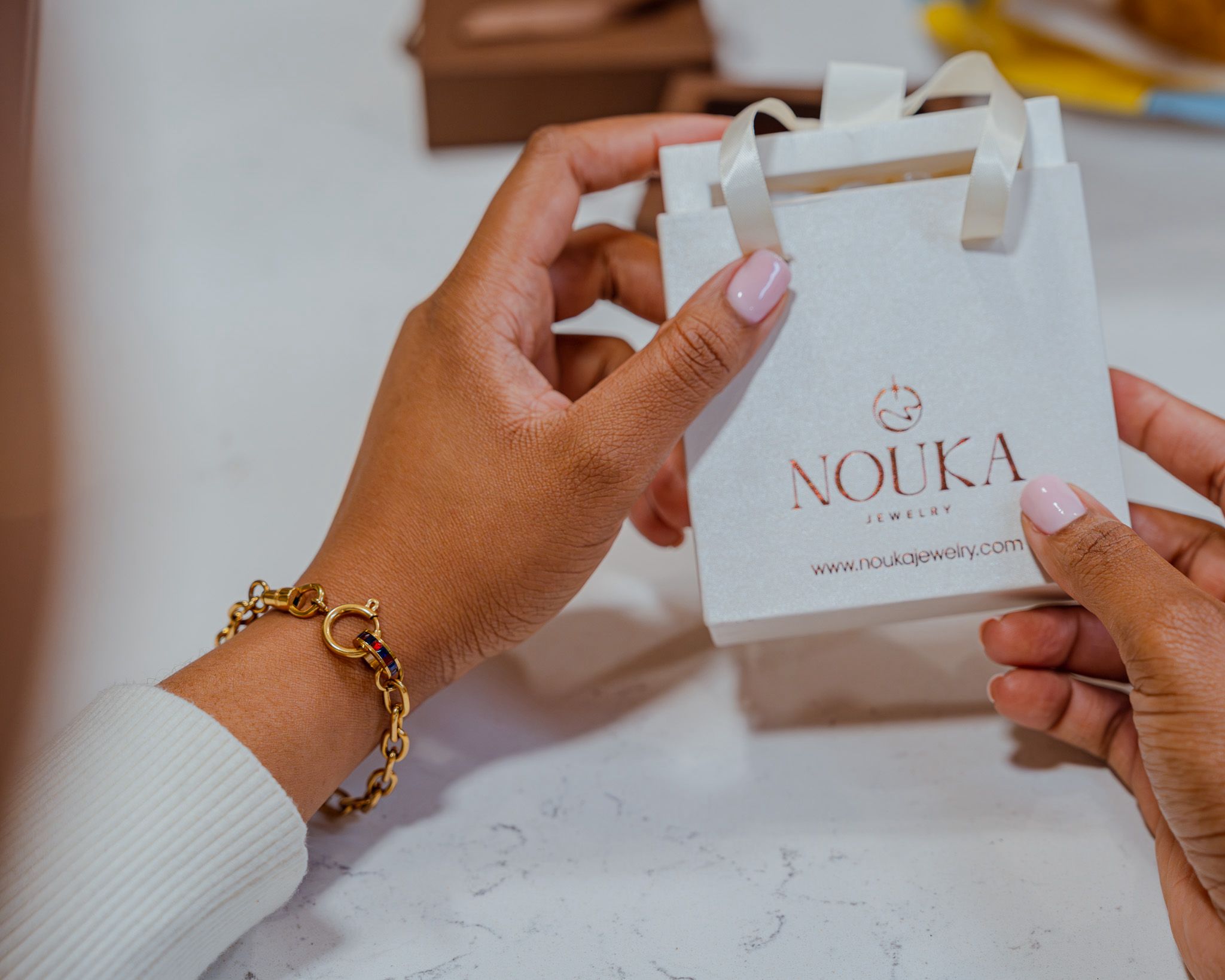 Person holding a white shopping bag with the logo 'NOUKA' and website, with another hand touching the bag.
