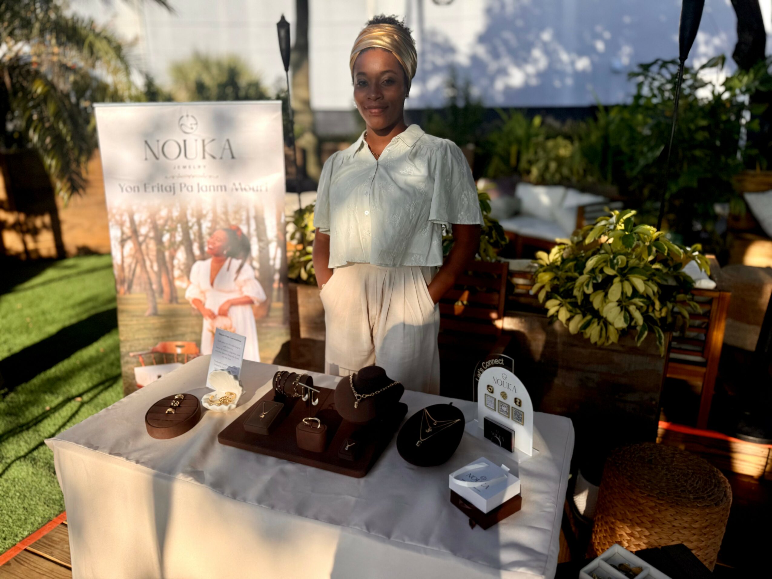 Woman standing behind a table with jewelry and promotional materials outdoors, with plants and a promotional banner nearby.