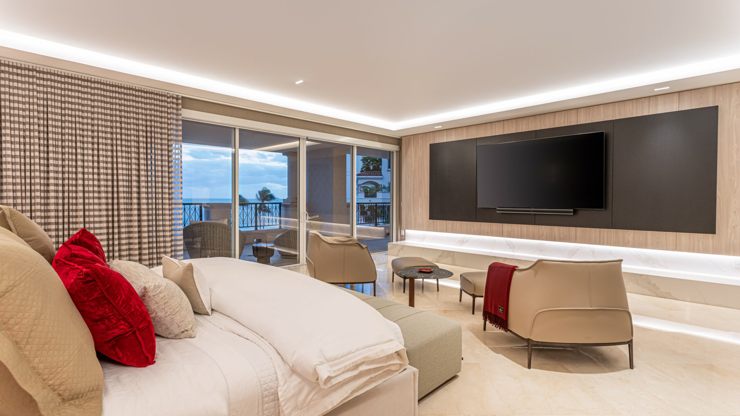 Living room with a large bed, red and white pillows, a TV on the wall, and a seating area near windows.
