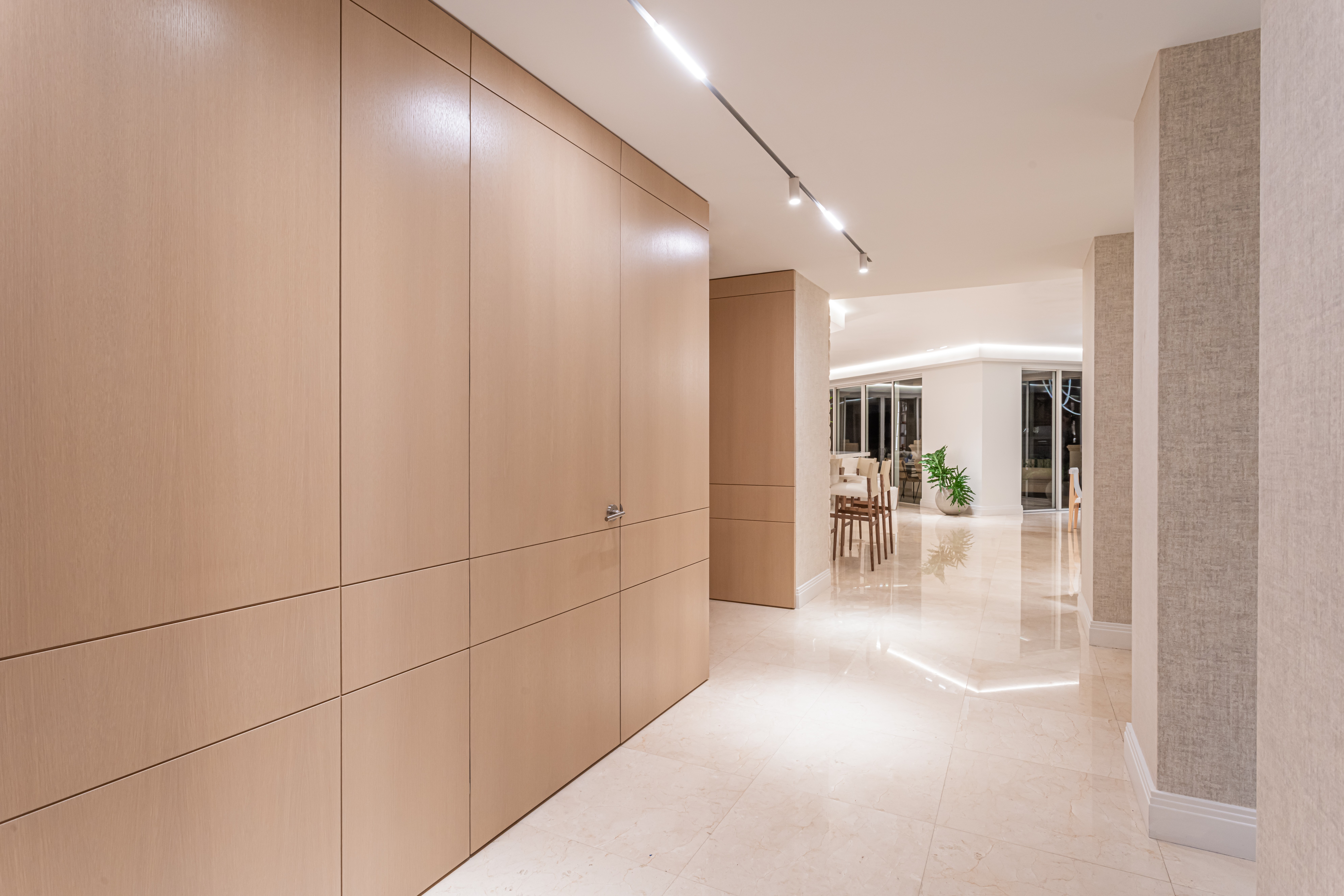 Modern interior hallway with wooden wall panels, bright lighting, and a view into a room with chairs and plants.