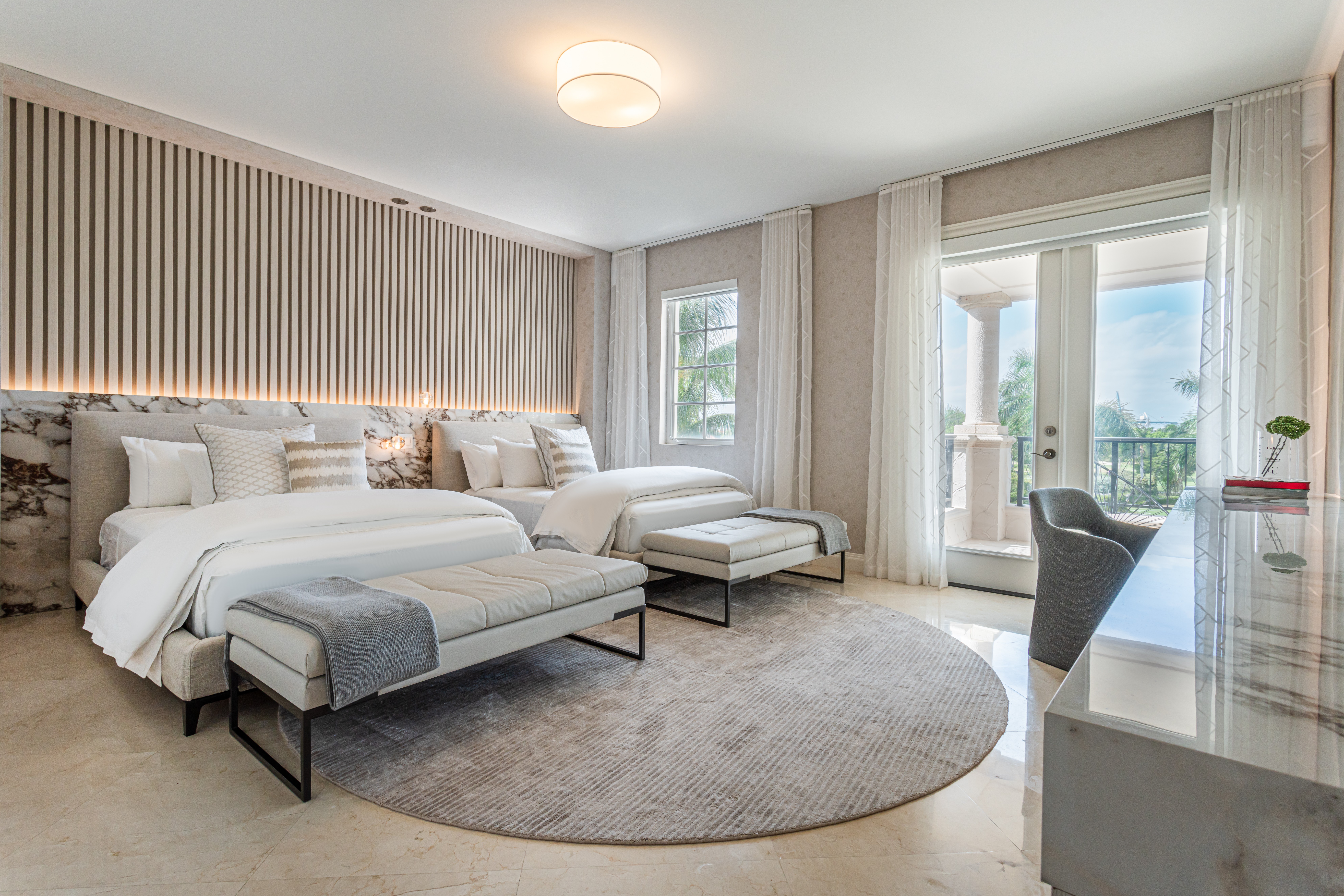 Bedroom with large bed, two benches, round rug, desk, and balcony door with curtains.