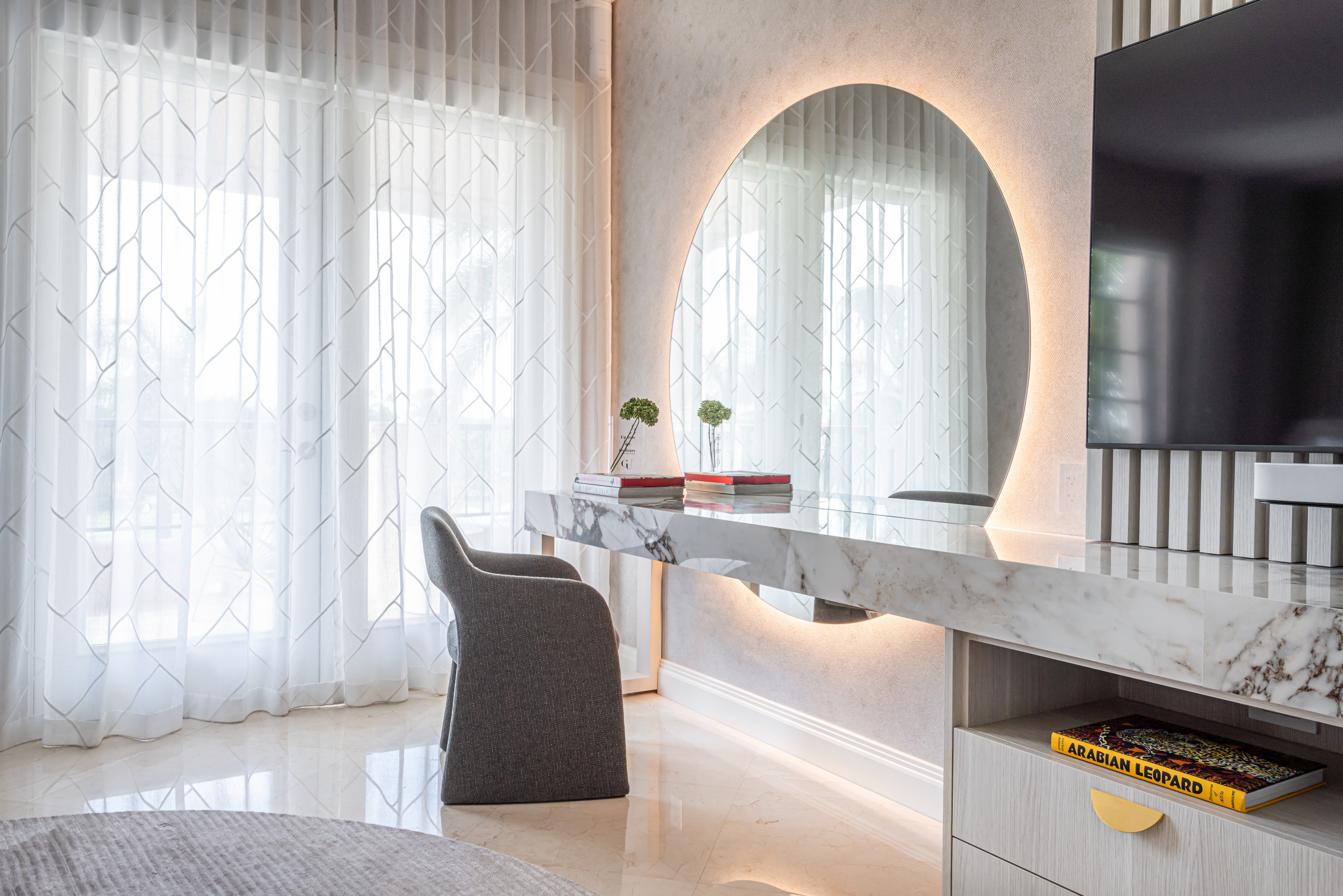 Modern interior with a marble desk, a mirror with backlit lighting, and a chair near large windows with sheer curtains.
