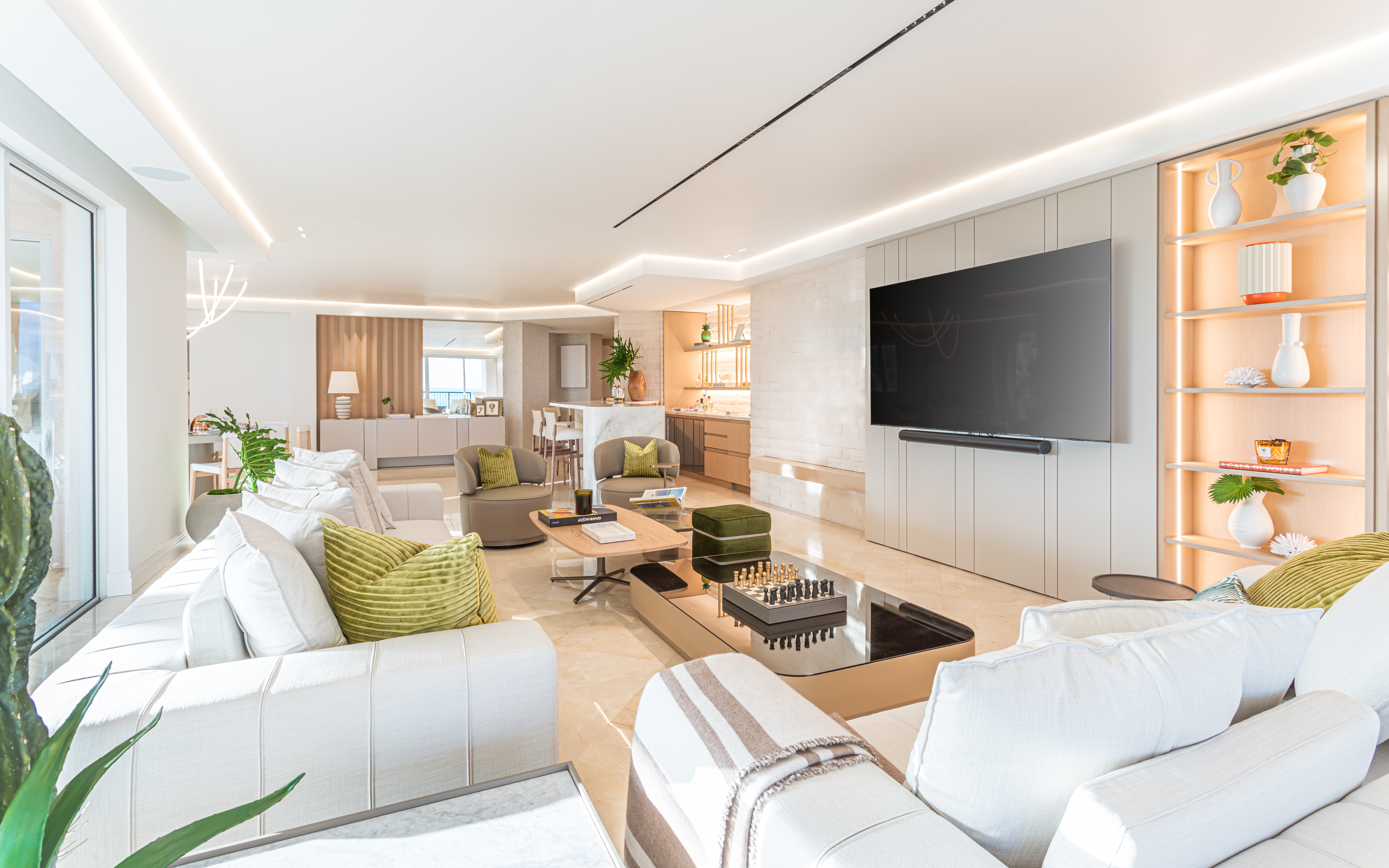 Modern living room with white sofas, a large wall-mounted TV, wooden shelves, and decorative plants.