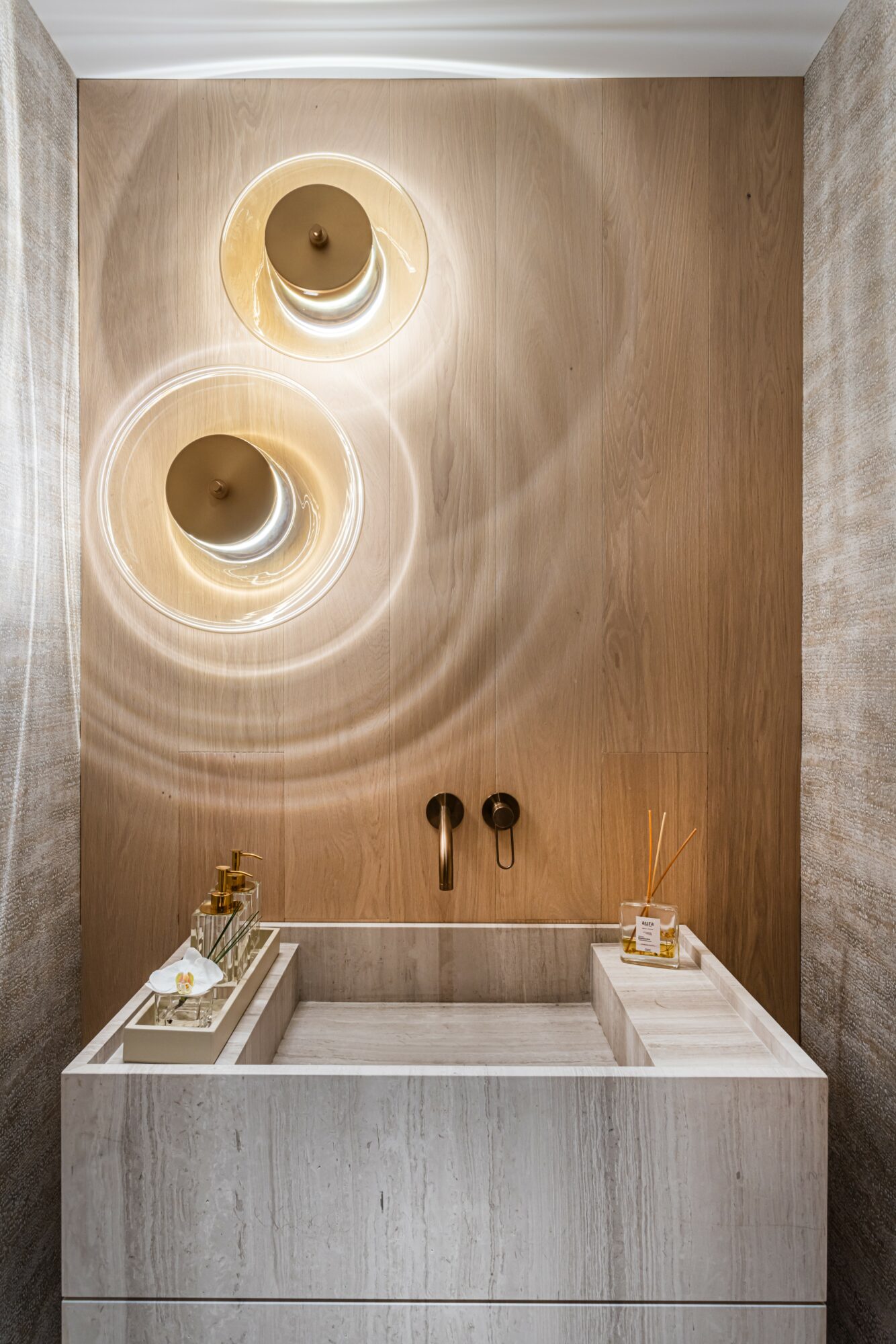 Bathroom sink with two black faucets, beige wall with two circular light fixtures, and decorative items on sides.