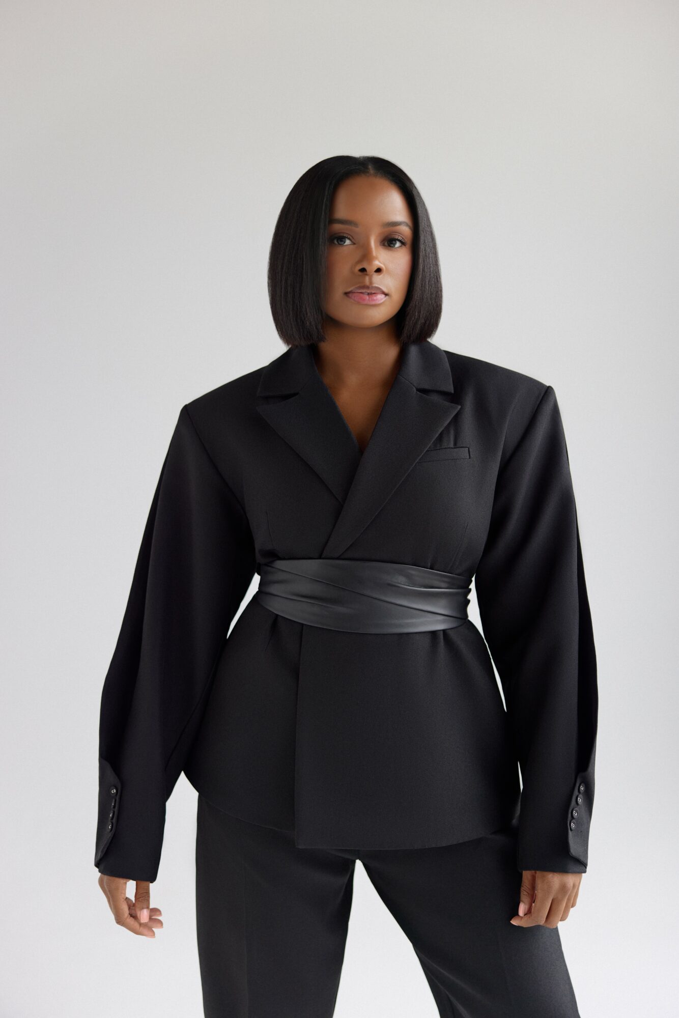 Woman in black suit with wide belt, standing against plain background.