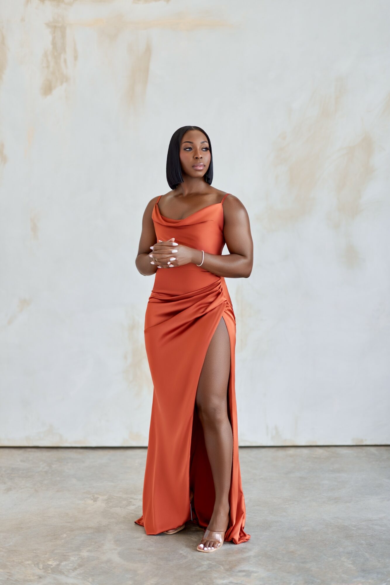 Woman in an orange dress with a slit, standing against a plain wall, looking to the side.