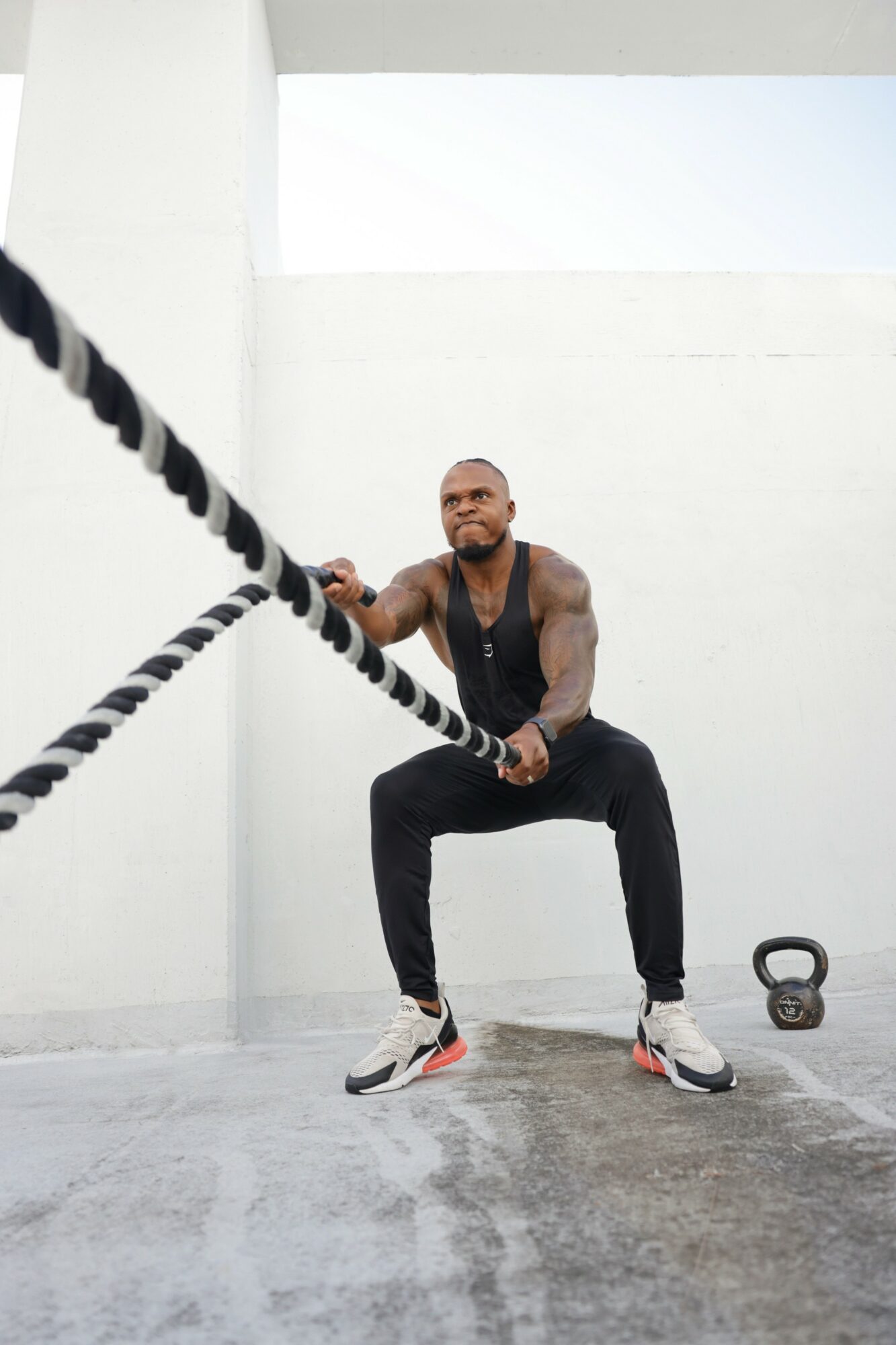 Man in black workout clothes holding a thick rope in a minimalistic room with a kettlebell nearby.