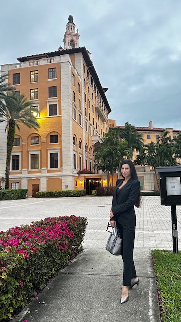 Woman in black suit holding a bag standing on sidewalk with building and trees in background.