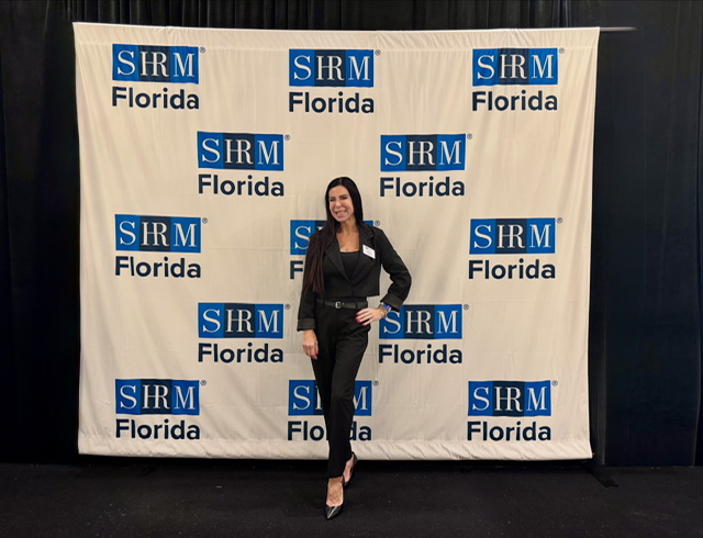 Woman in black suit standing in front of a backdrop with SRM Florida logos, smiling, with dark hair and heels.