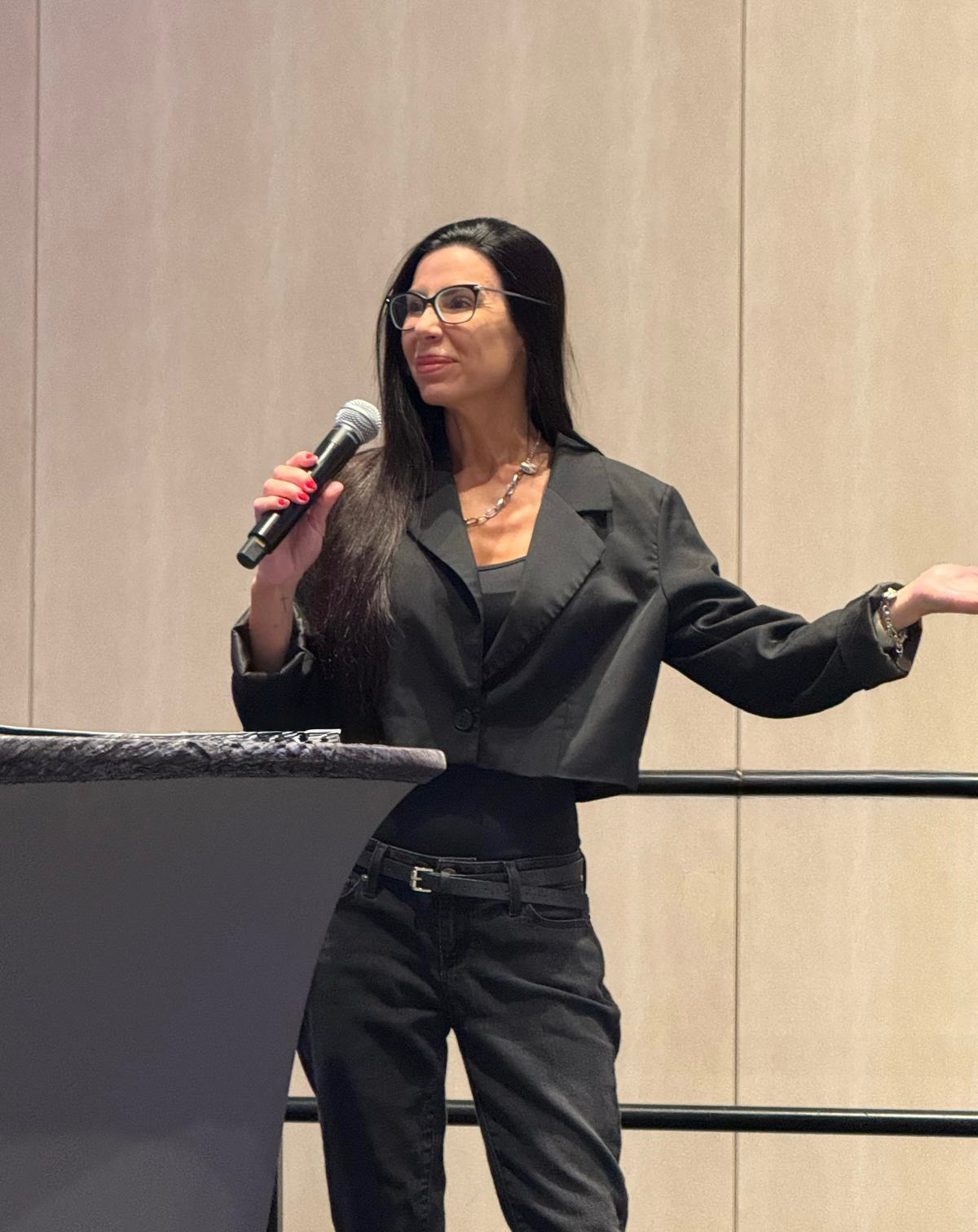Woman with long dark hair and glasses speaking into a microphone, gesturing with her left hand, wearing a black blazer and pants.