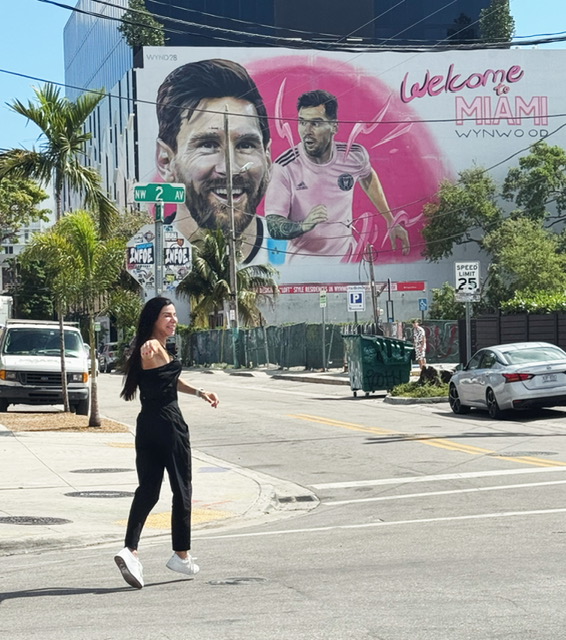 Woman walking across a street with a large Miami welcome billboard in the background.