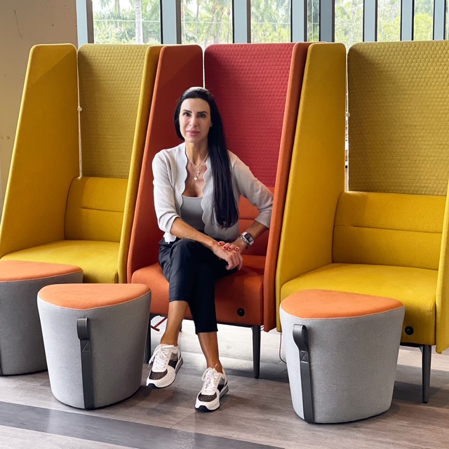 Woman with long dark hair sitting in a colorful high-backed chair, surrounded by yellow and red chairs, in a modern indoor space.
