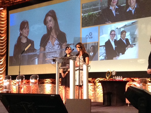 Two women stand at a podium on stage, with large screens showing their images behind them.