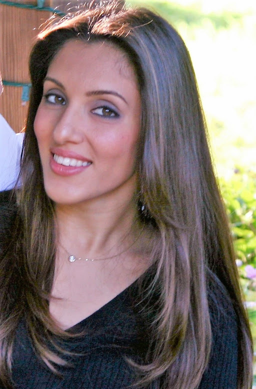 Young woman with long brown hair smiling outdoors, wearing a black top and a necklace.