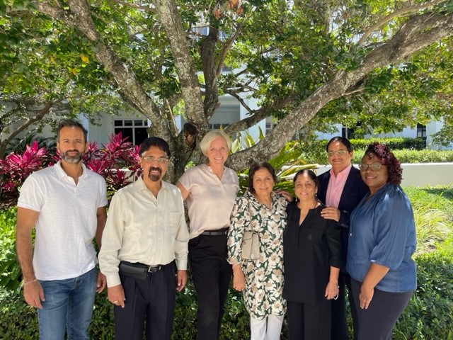 Seven people standing outdoors under a large tree with green leaves and pink flowers, smiling at the camera.
