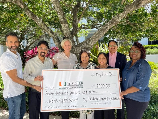 Group of seven people standing outdoors holding a large check under a tree with green leaves.