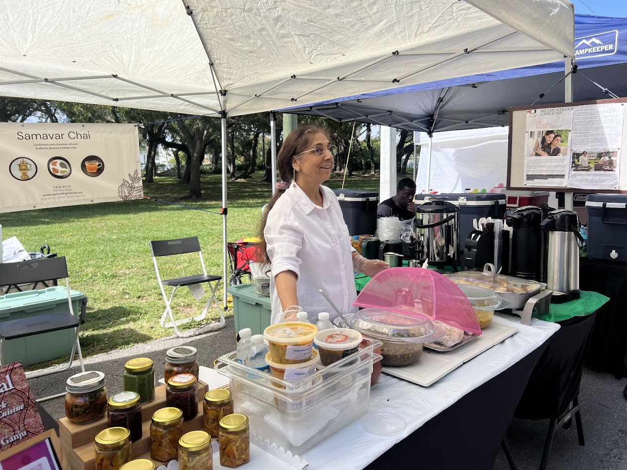 Woman standing behind a table with jars, containers, and a pink bag at an outdoor market stall under a canopy.