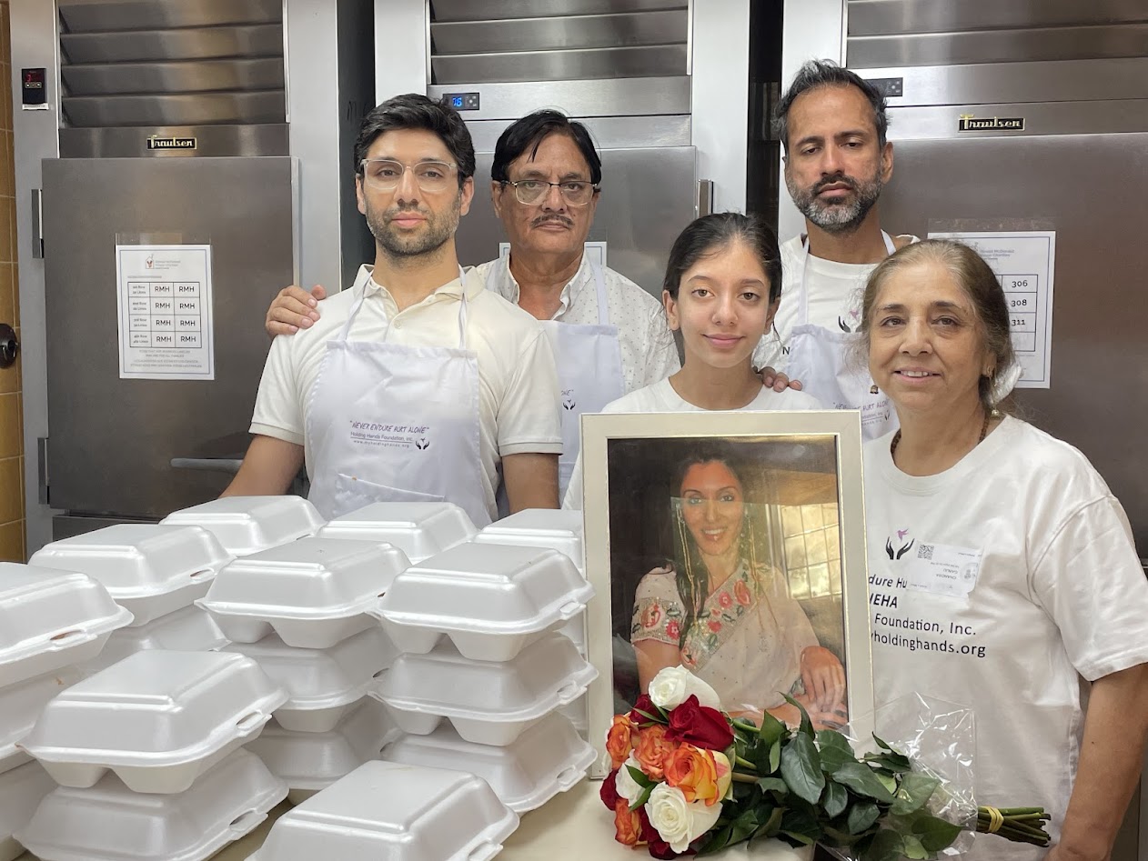 Group of five people standing behind stacked white food containers, with a framed photo and flowers in front, in a kitchen.