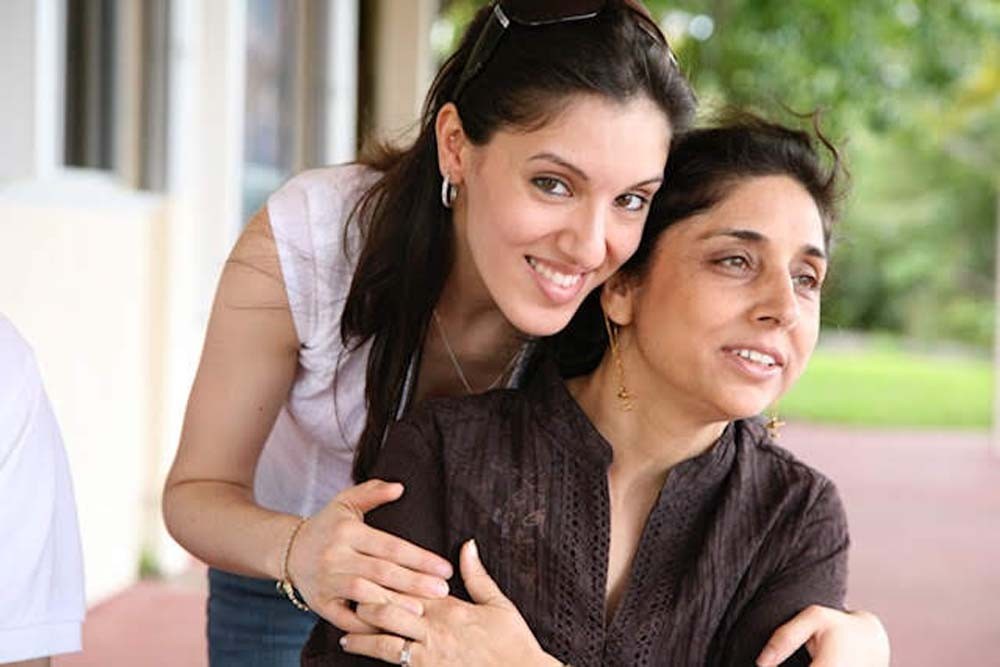 Two women sitting outdoors, one smiling and leaning on the other, with greenery in the background.