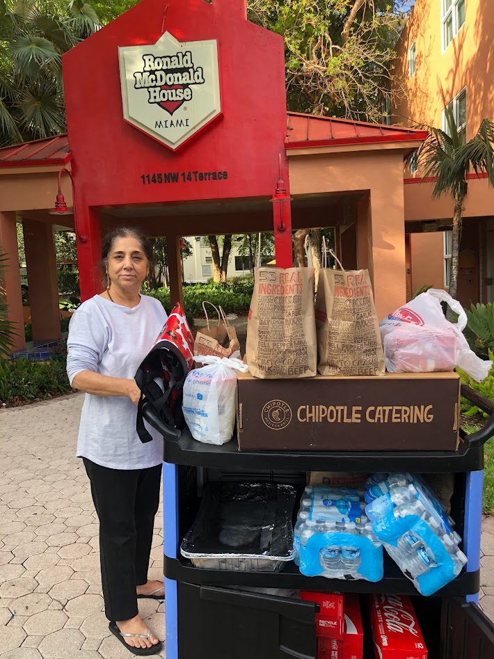 Woman standing outdoors next to a cart with food and beverage supplies, in front of a Ronald McDonald House sign.