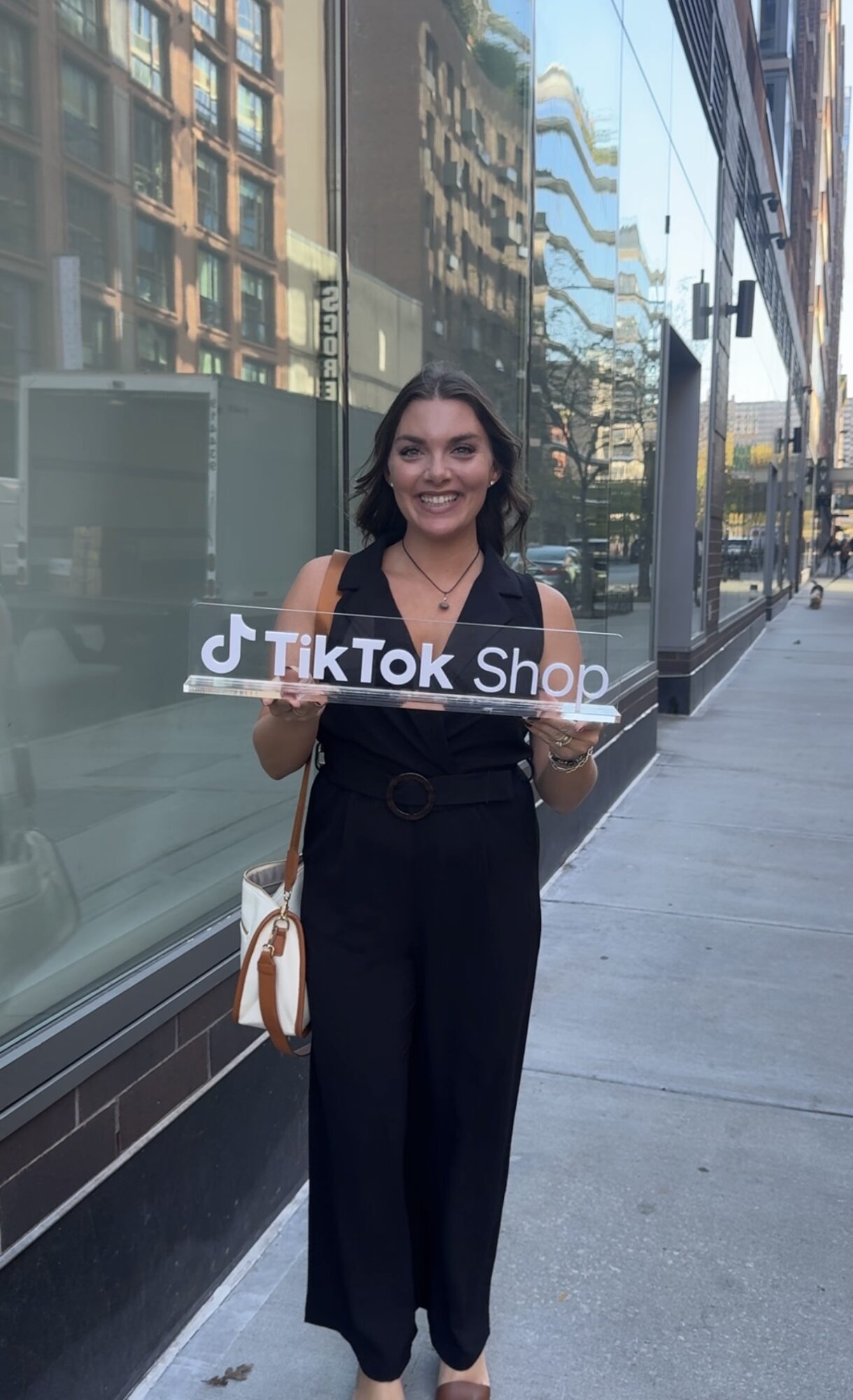 Young woman with dark hair smiling, holding a TikTok Shop sign, standing on city sidewalk near glass building.