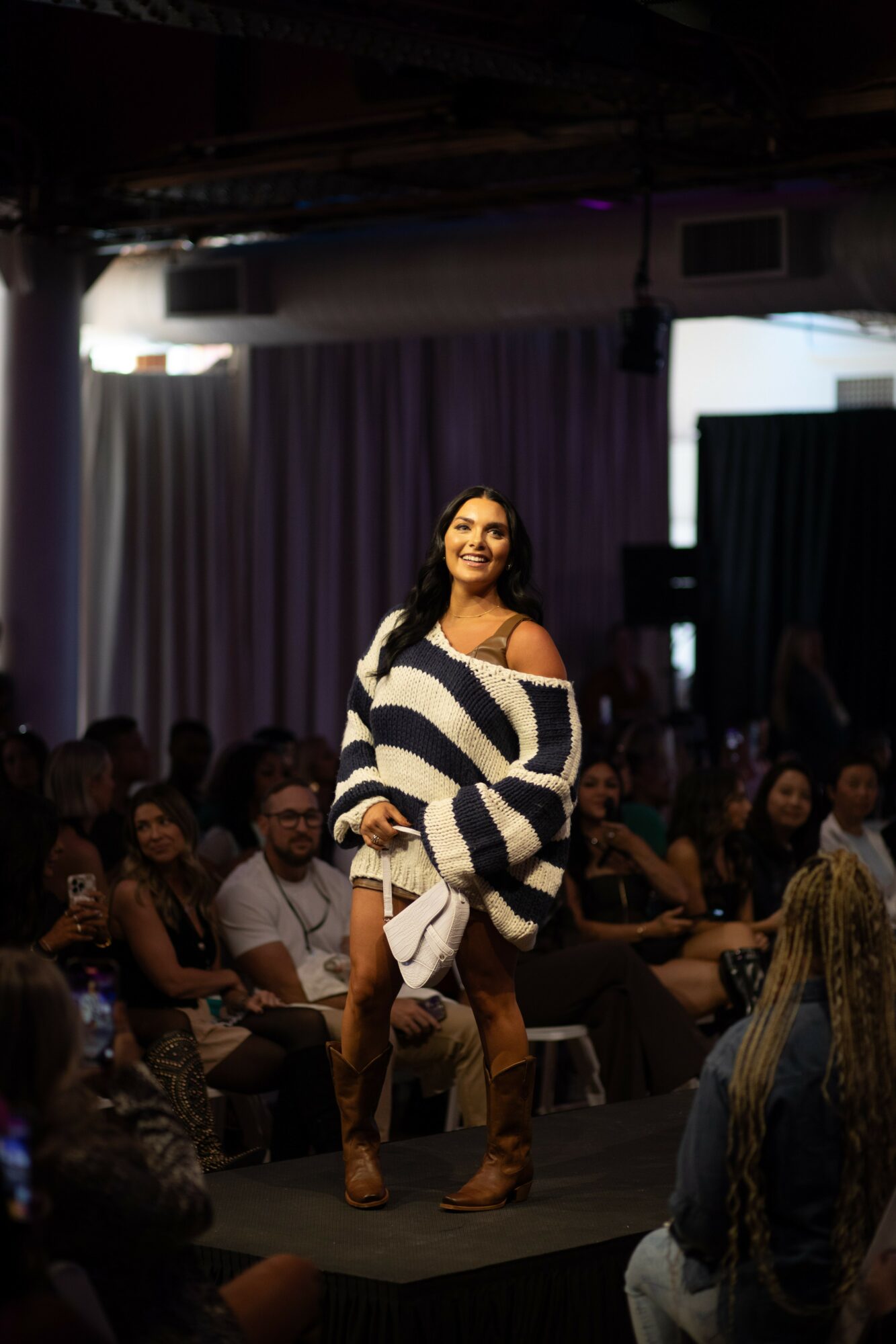 Woman walking on stage in front of seated audience, smiling, wearing a striped sweater and shorts.