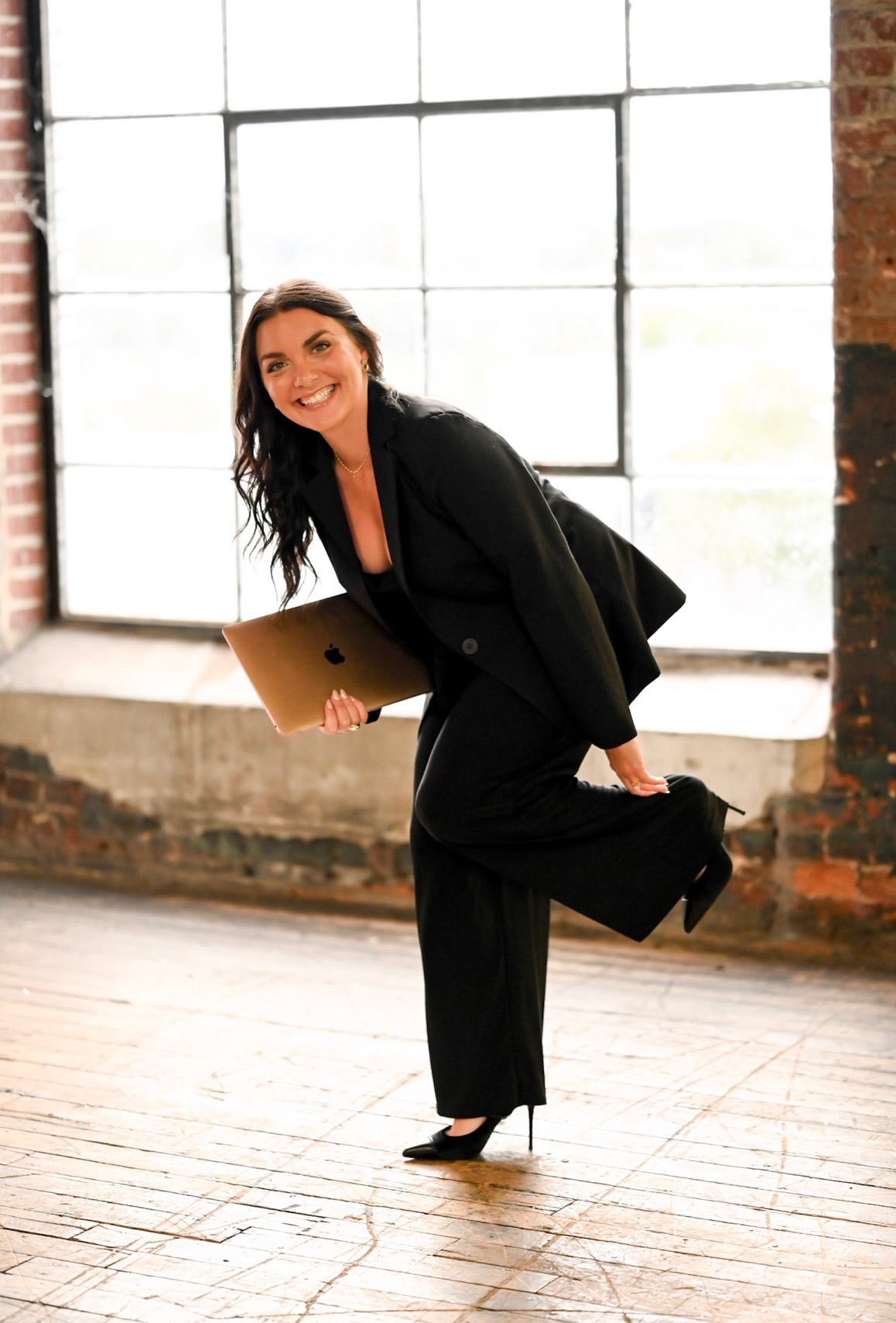Woman in black suit holding a folder, smiling, standing indoors near large window with brick wall background.