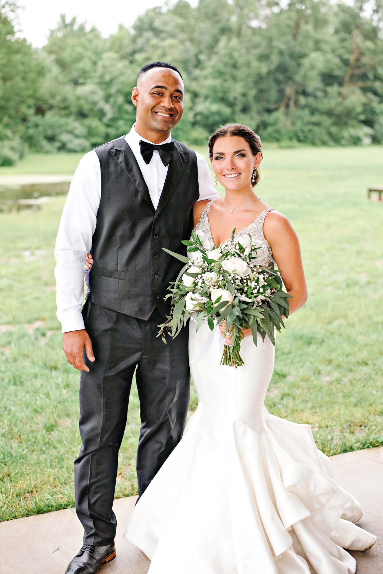 A bride and groom standing outdoors on grass, smiling, with trees in the background. The bride holds a large bouquet.