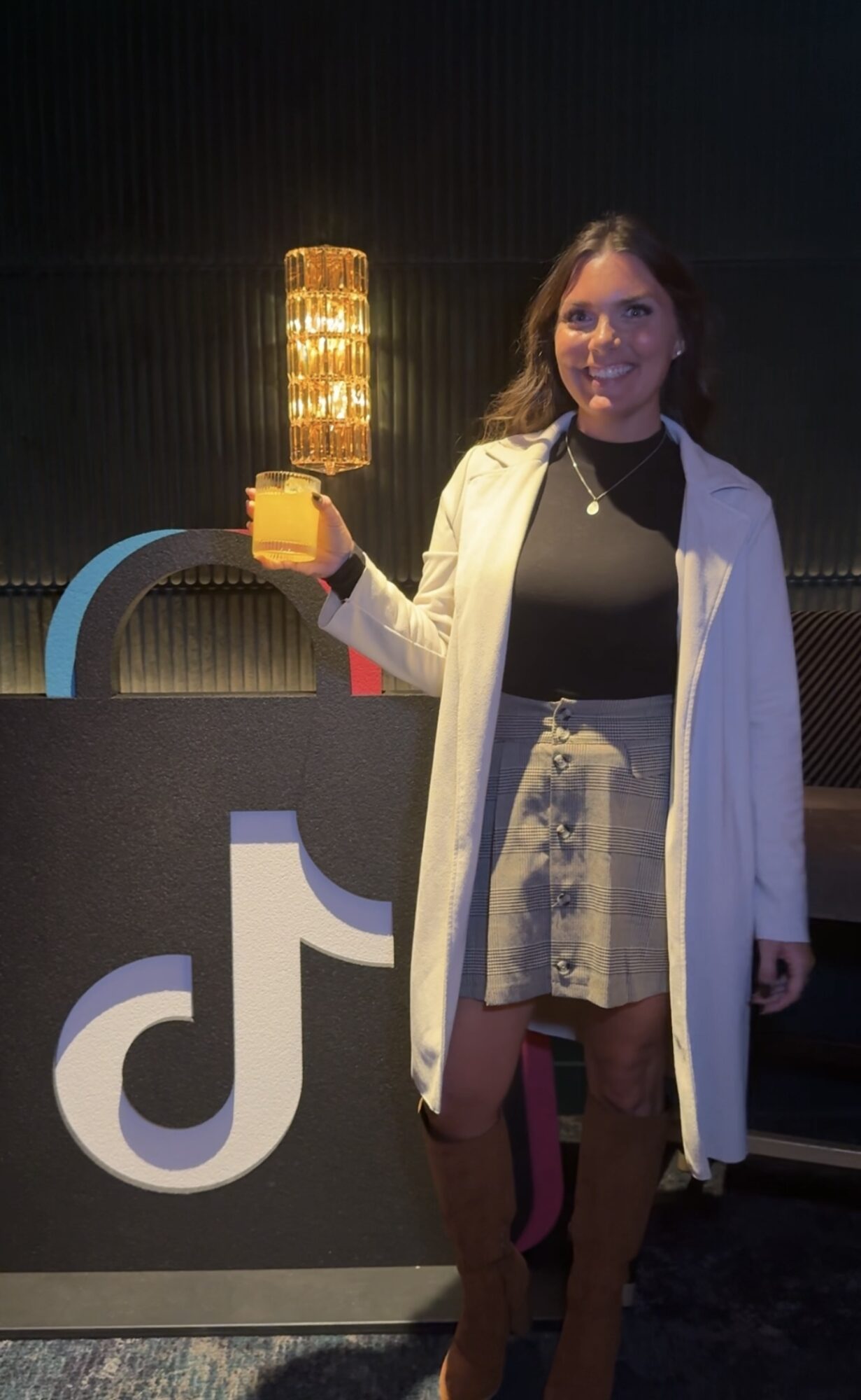 Woman standing next to a TikTok logo display, holding a yellow object, smiling, in a dark room with a lamp behind her.
