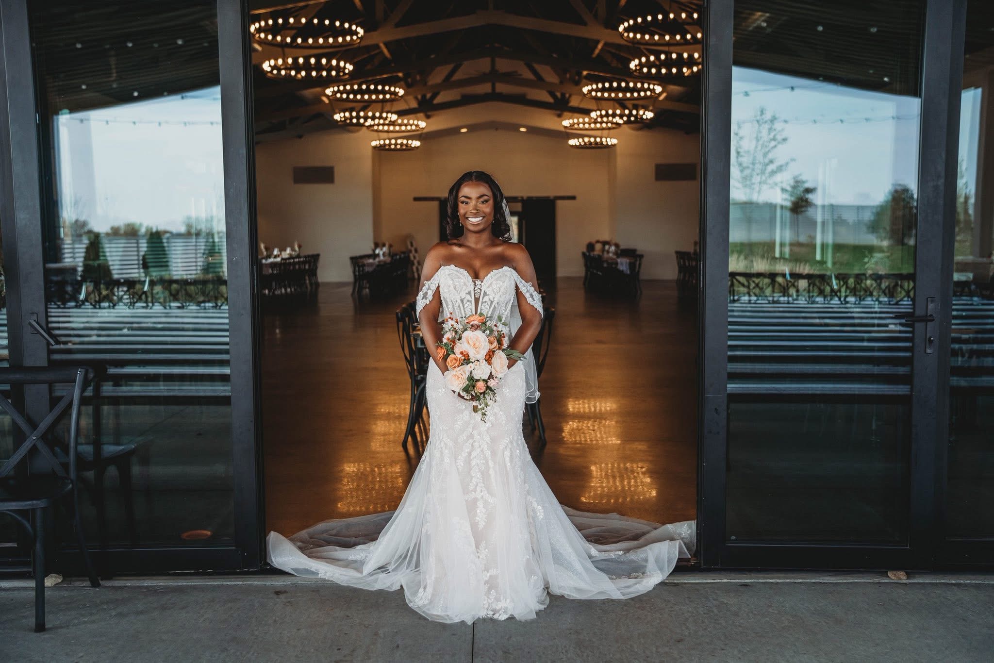 Bride in a white wedding dress holding a bouquet, standing in a decorated hall with large glass doors open behind her.