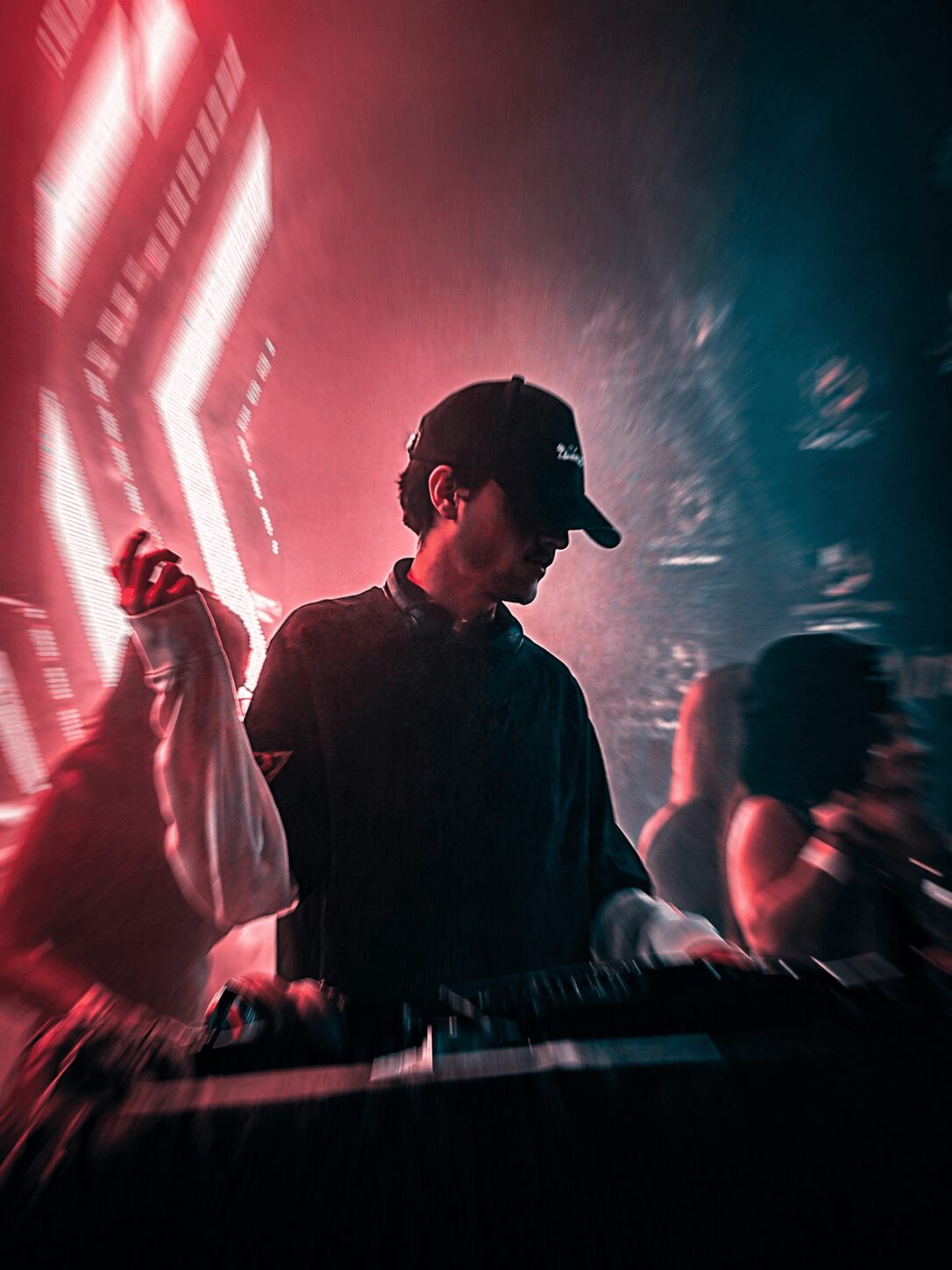 Person wearing a cap and dark clothing, standing in a dimly lit environment with colorful lights and other people nearby.