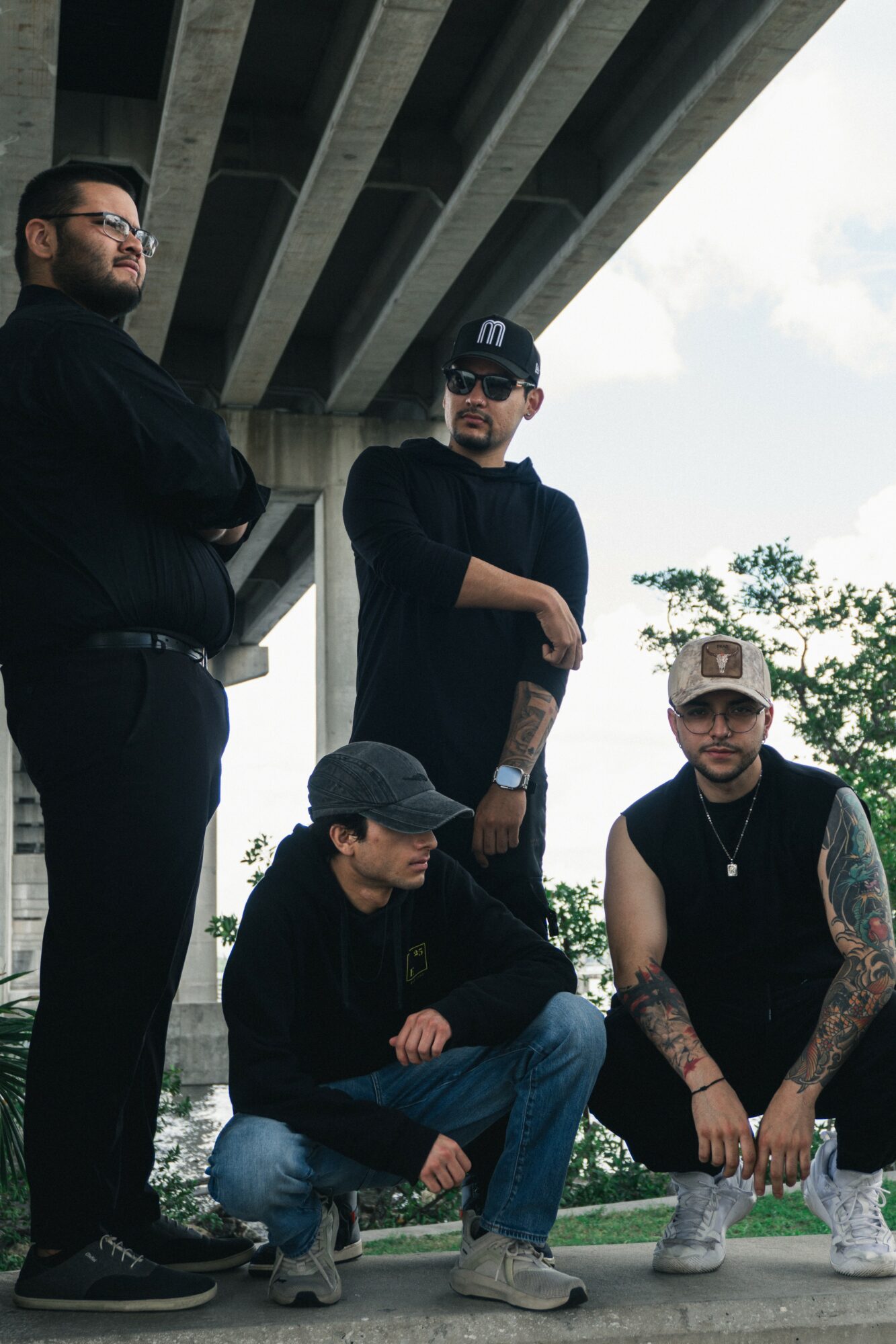 Four men under a bridge, two standing and two sitting, with trees and sky in background.